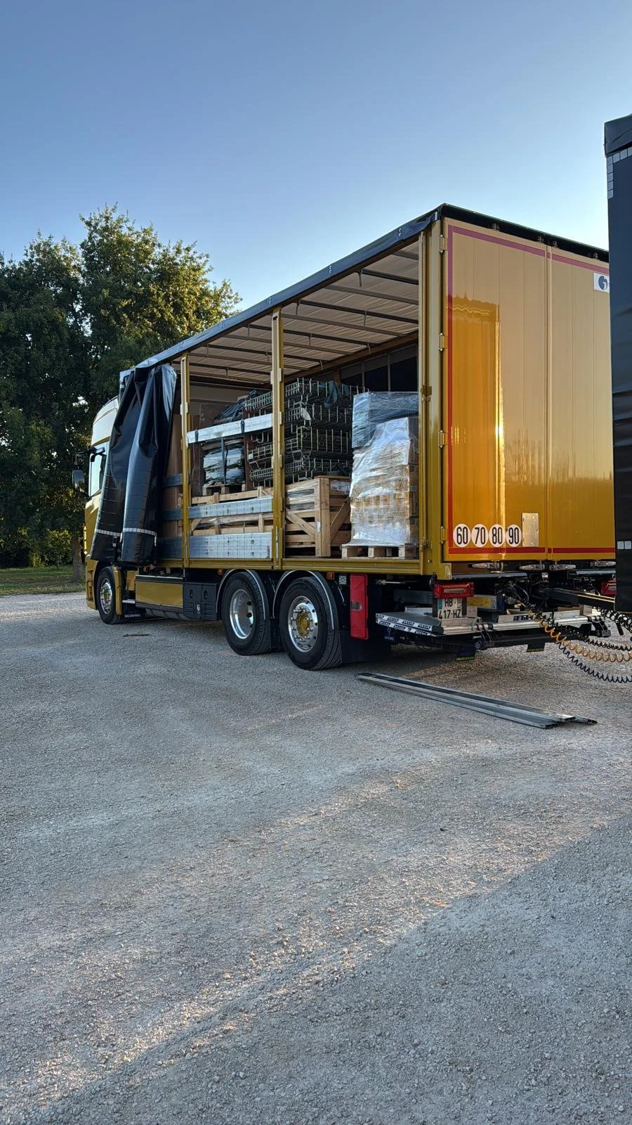 Un camion de transport avec une remorque ouverte par le côté contenant des palettes, des étagères métalliques et divers objets encadrés de bois dans un espace extérieur.