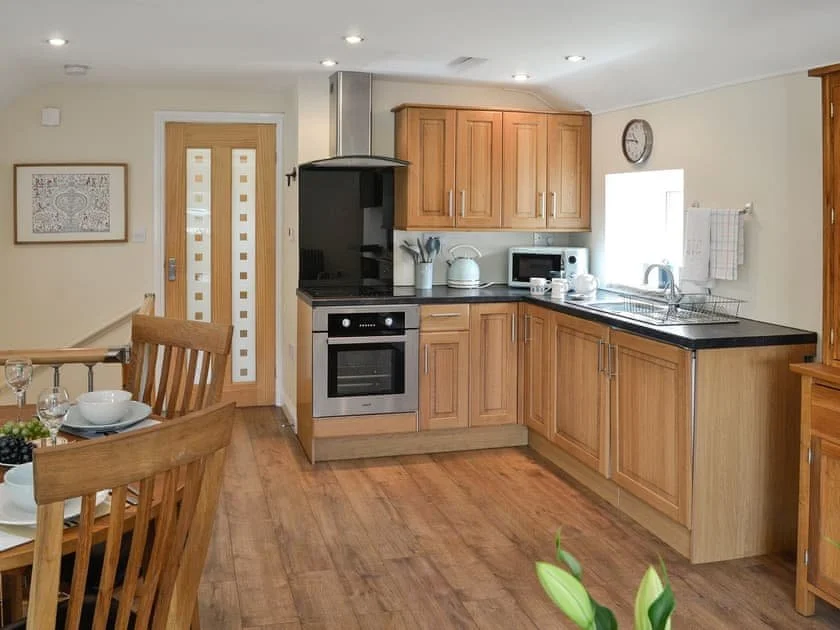 Kitchen with wooden cabinets, black countertop, stainless steel oven and microwave, white kettle, and dish drying rack. There is a clock on the wall and a window with a towel hanging. Dining table with chairs and place settings are visible in the foreground.