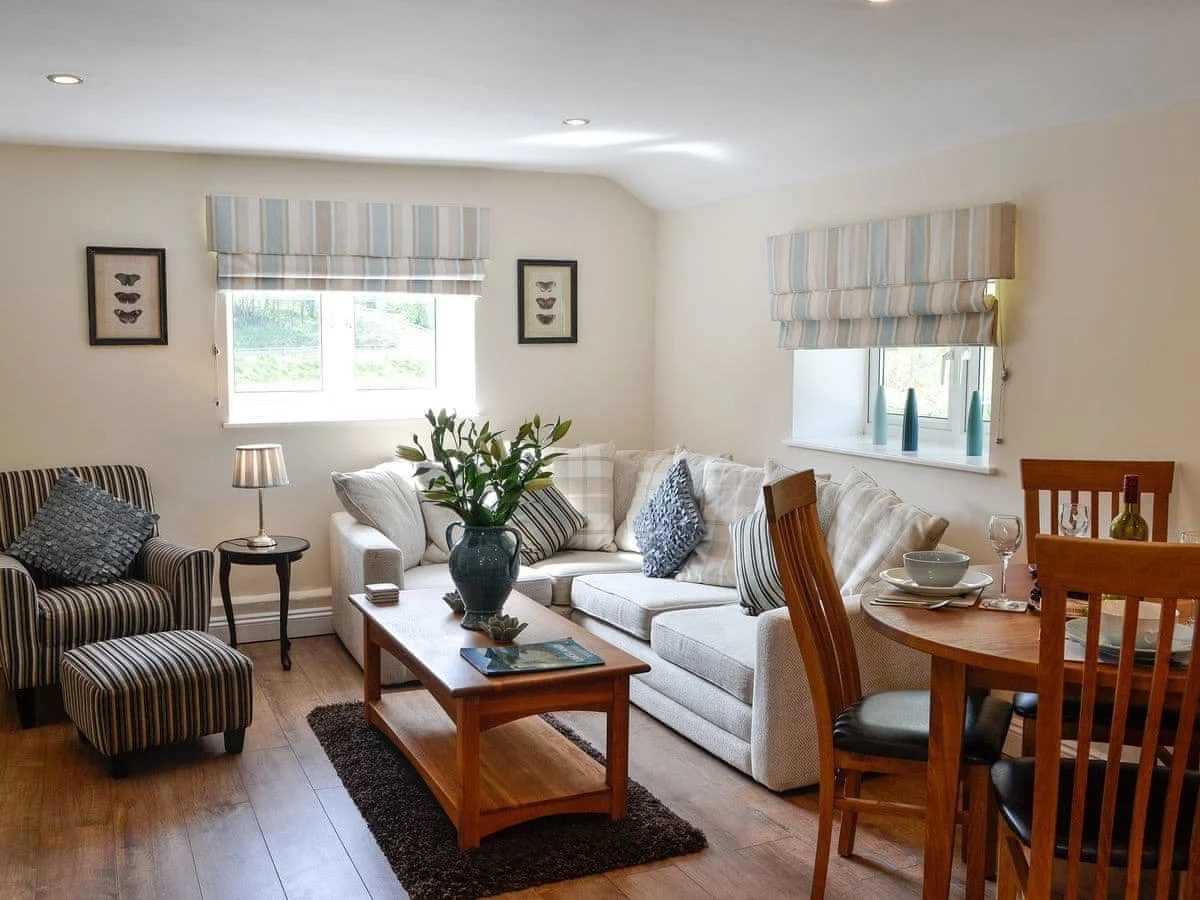 Living room with beige sofa, striped armchair, wooden coffee table with vase, and dining area with wooden table and chairs. Windows have striped blinds, and decorative items include framed butterfly prints and vases.