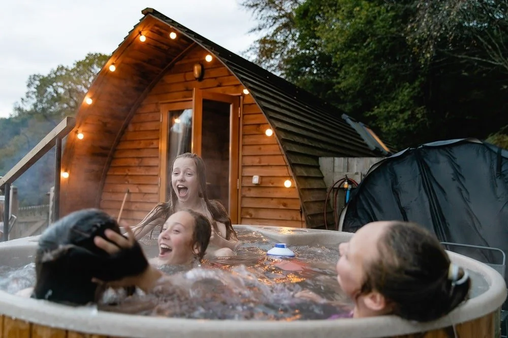 Four children enjoying a hot tub outdoors in front of a wooden cabin decorated with string lights during evening.