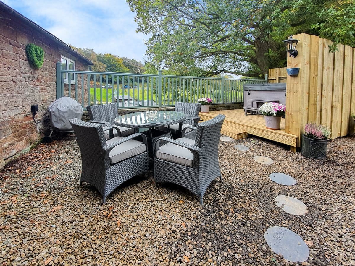 Backyard patio with a round glass table and four wicker chairs with cushions, surrounded by gravel, potted flowers, and a wooden deck with fencing and trees in the background.