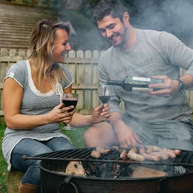 A woman and a man grilling meat outdoors, smiling and enjoying each other's company.