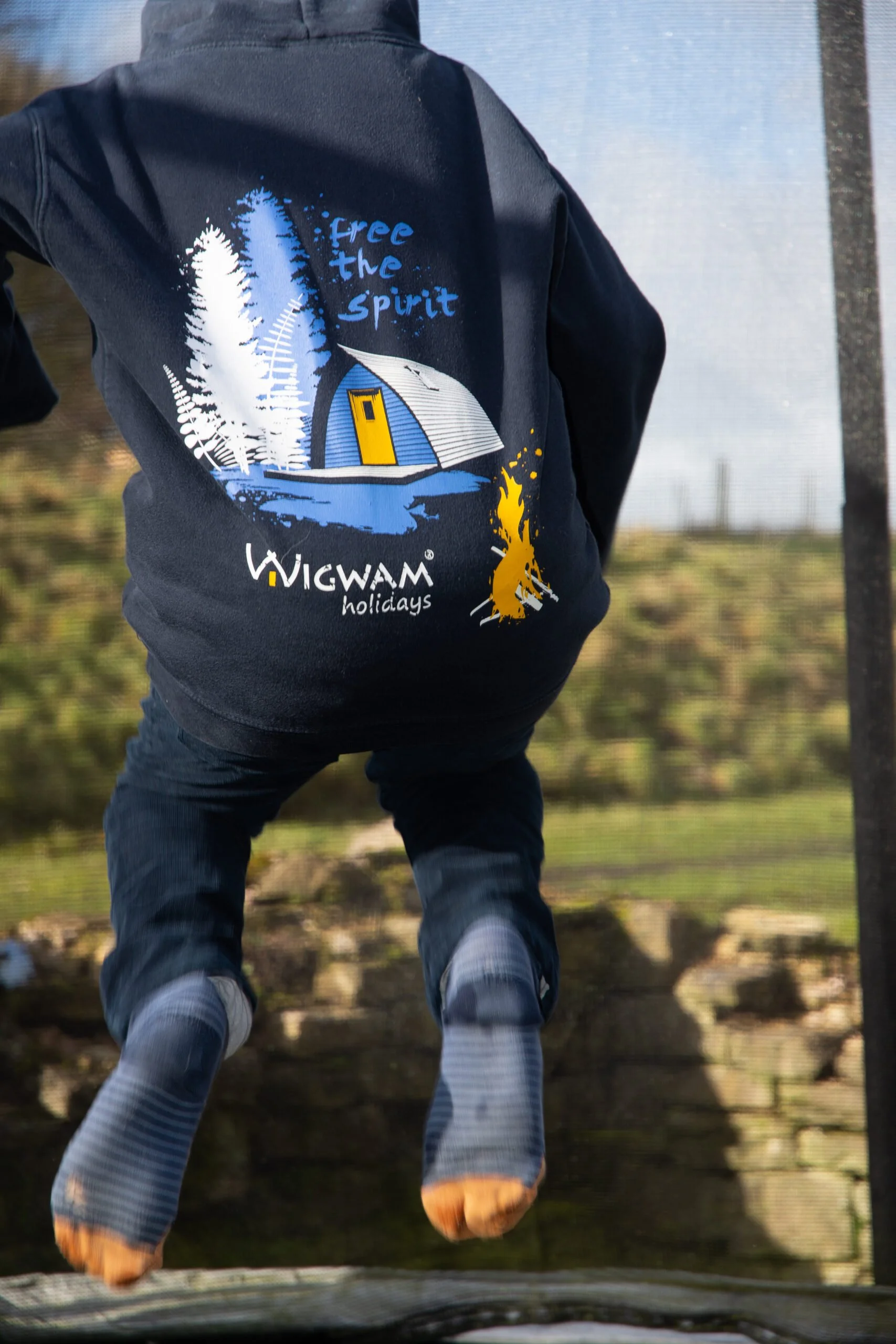 Person in a black hoodie with a jacket featuring a design of trees, a tent, and the words "free the spirit" and "WIGWAM holidays." The person is jumping off a platform outdoors, with blurred trees and sky in the background.