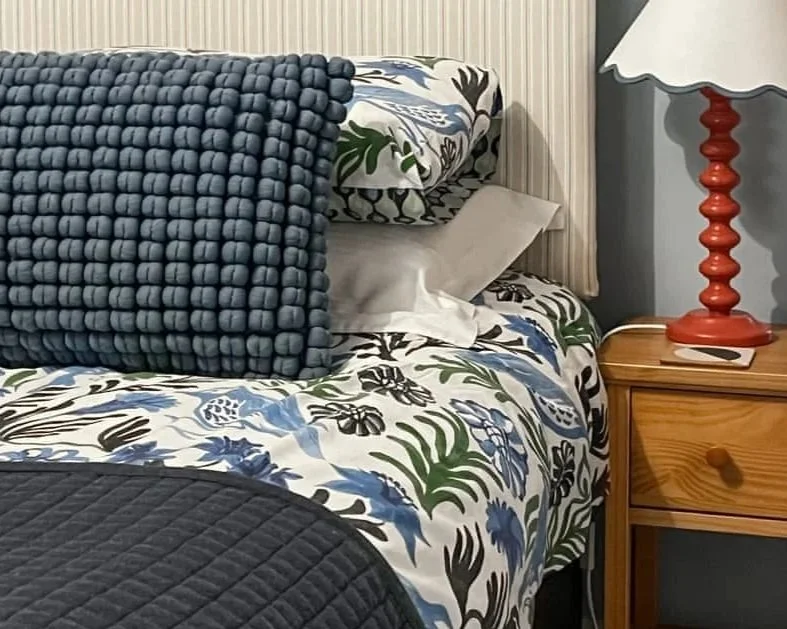 A bed with a floral patterned comforter, white and patterned pillows, a textured blue throw blanket, and a navy quilted bedspread. To the right, a wooden nightstand holds a red lamp with a white shade.
