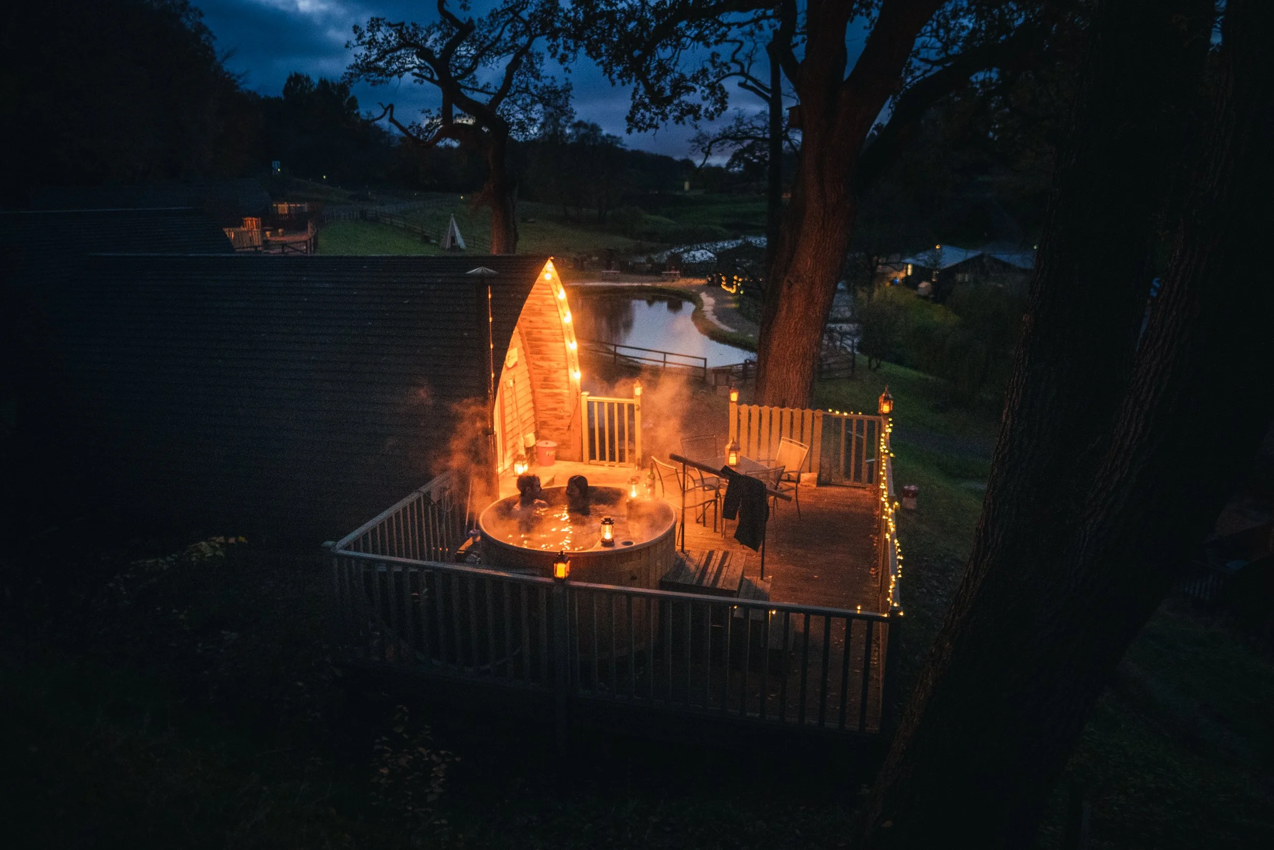 People enjoying a hot tub on a wooden deck illuminated by string lights, overlooking a calm river and trees at dusk.