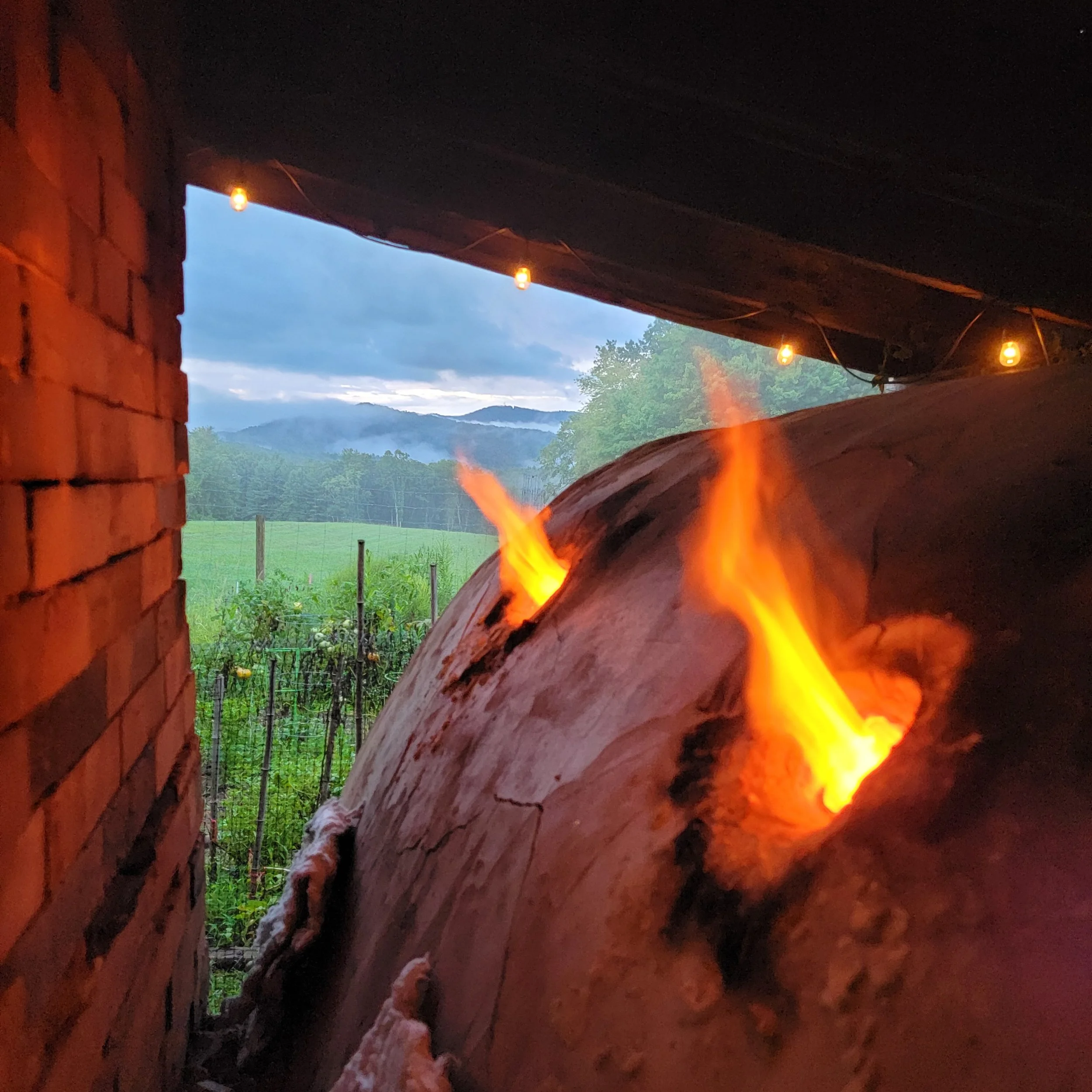 A close-up view of a flame form a wood kiln firing pottery, overlooking a green field and distant mountains at dusk with string lights hanging overhead.