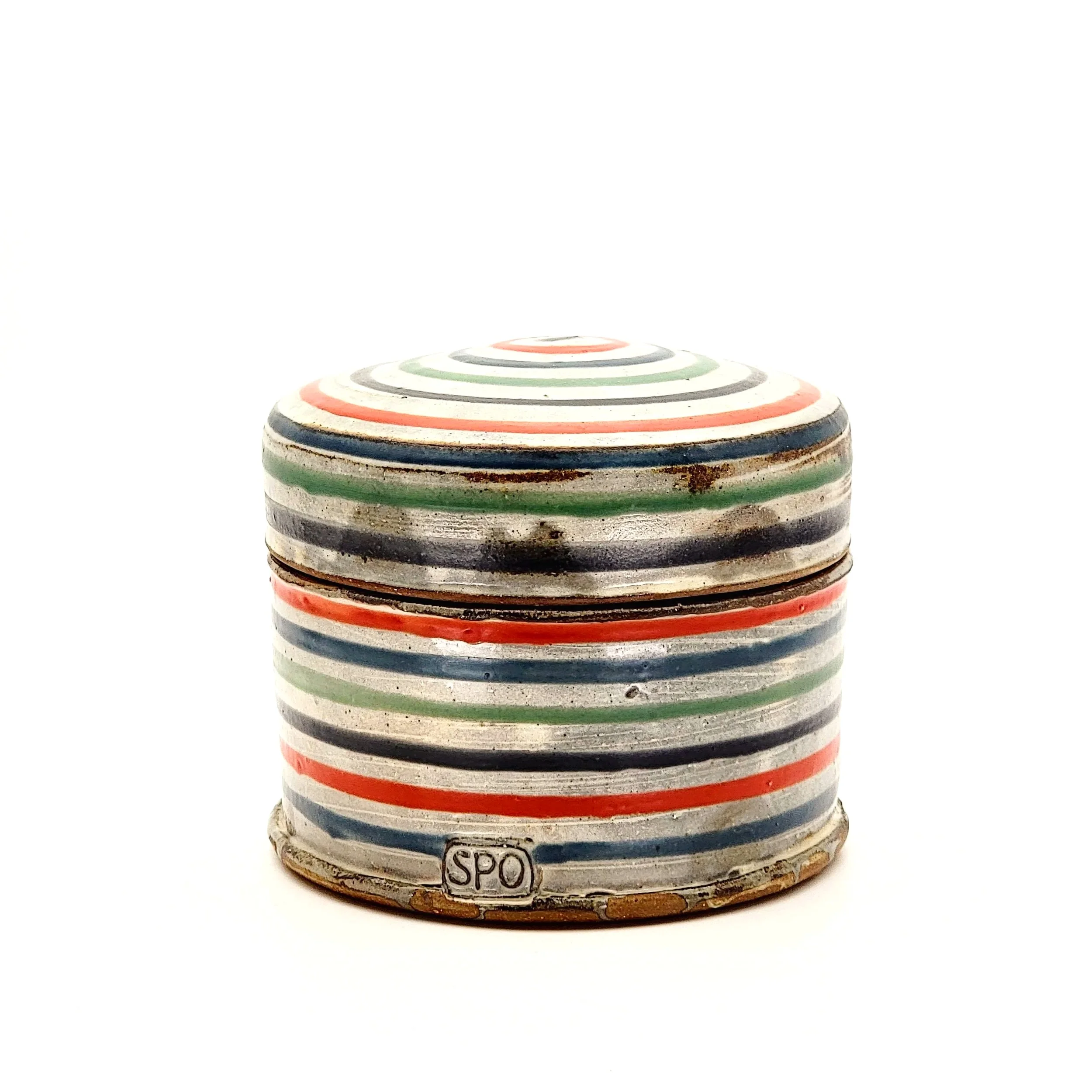 A vintage, round ceramic container with colorful concentric rings in red, green, black, and blue, featuring a lid with similar pattern, small brand mark 'SPO' at the base, and a weathered, rustic appearance.