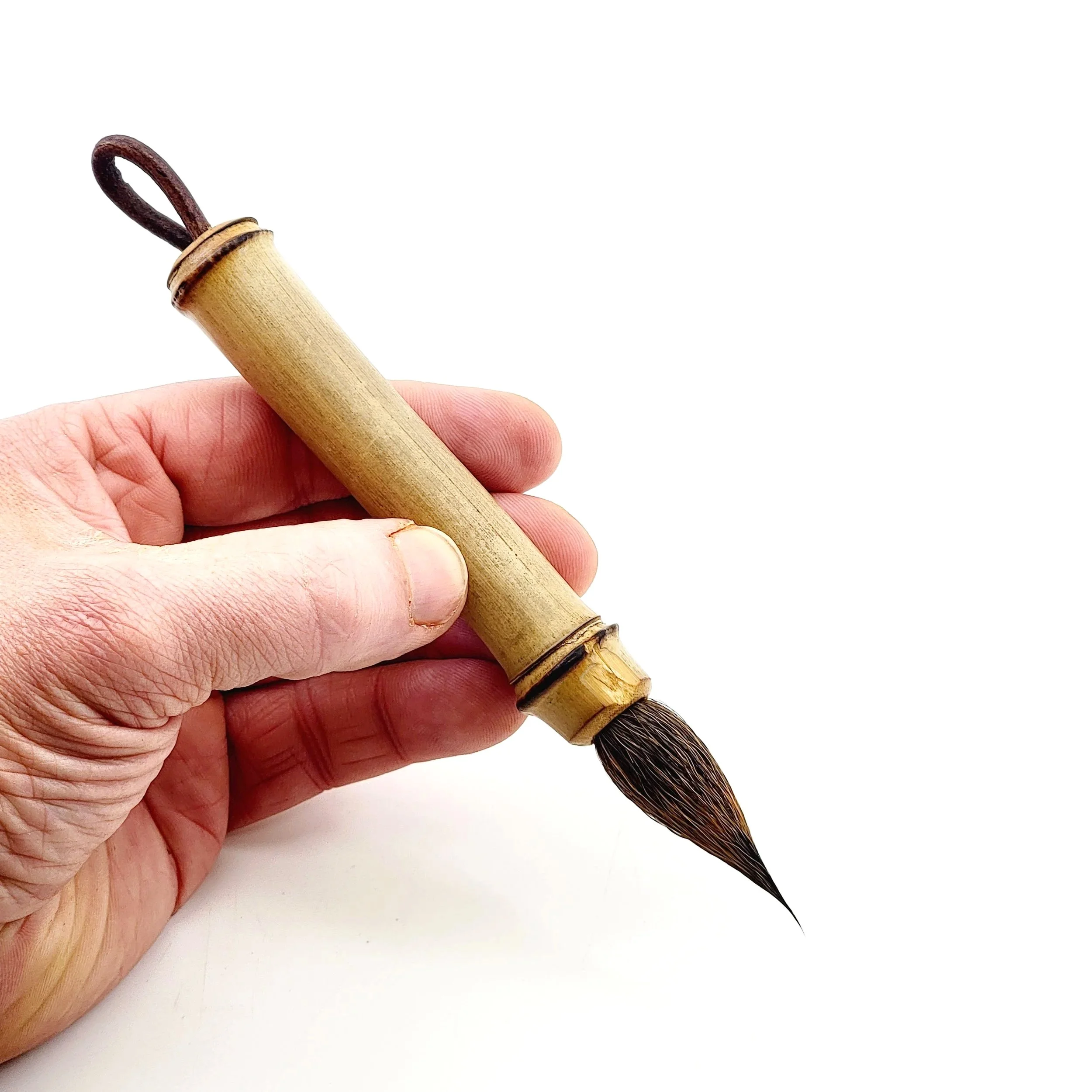 Hand holding a bamboo brush with dark bristles and a looped leather strap on the handle, on a white background.
