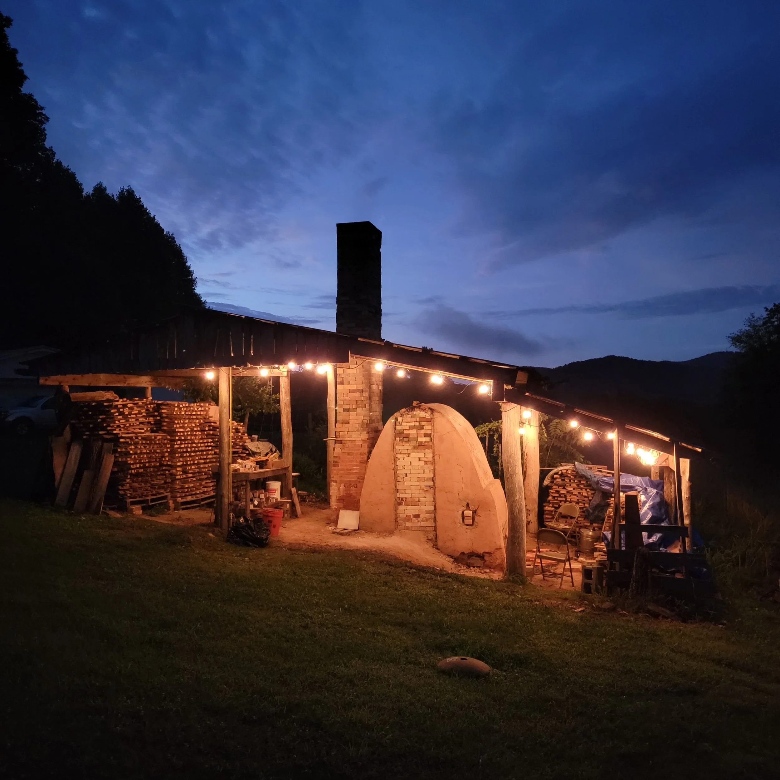 Outdoor wood-fired kiln under string lights at dusk, with stacked wood and outdoor chairs surrounding it, mountains in the background.