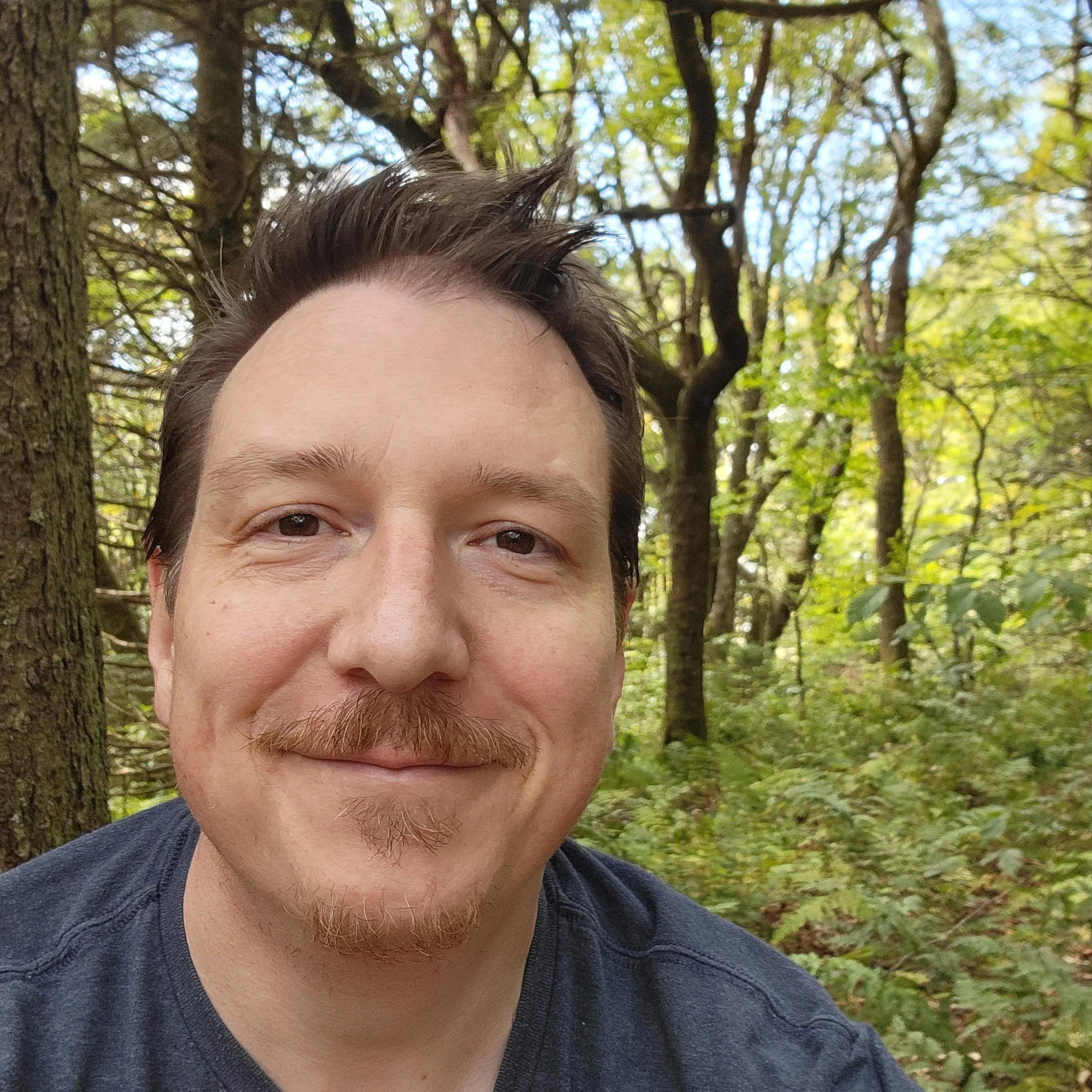 Artist Sean O'Connell smiling in a forest with trees and green foliage.