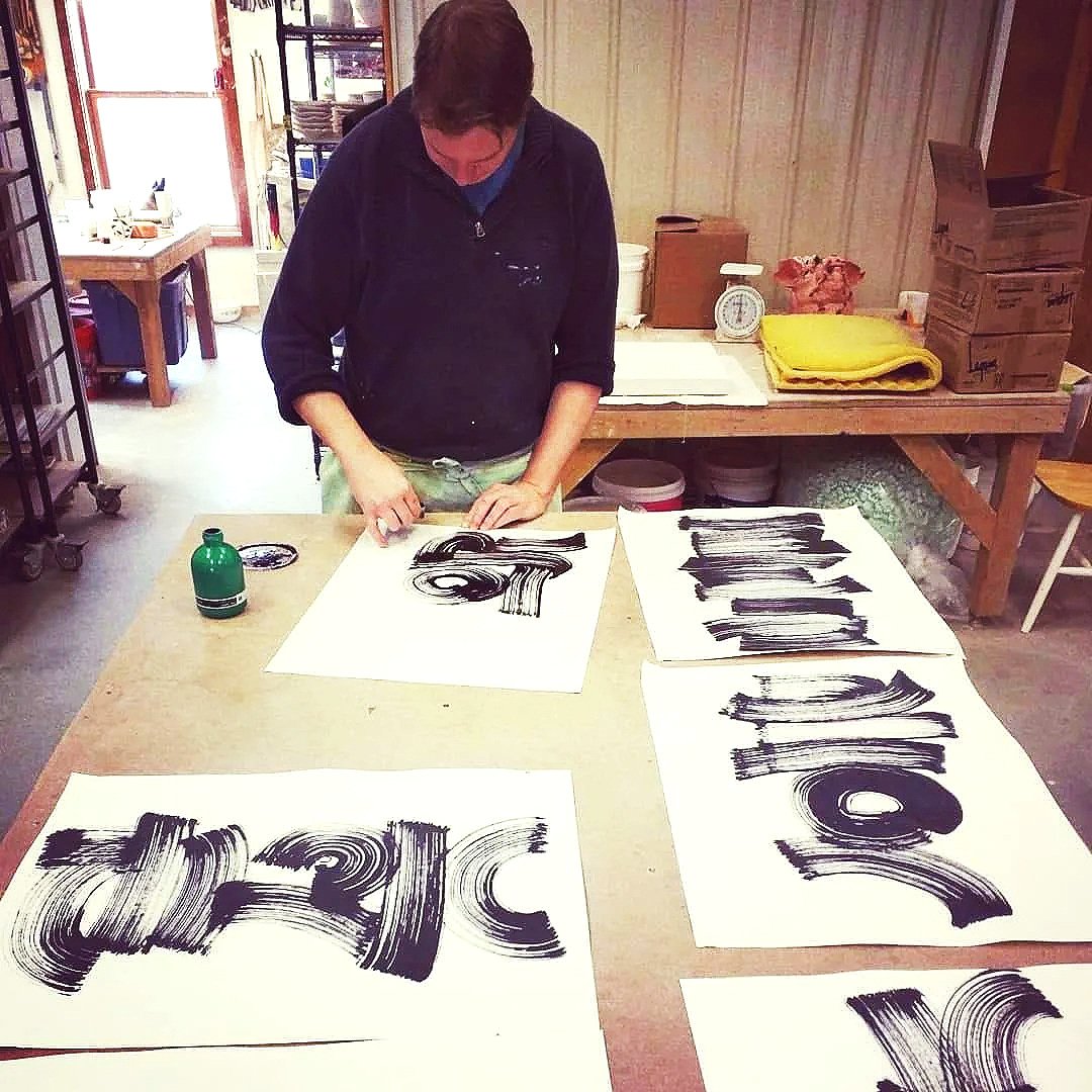 An artist is working on large abstract black brushstroke paintings on white paper, arranged on a wooden table in a well-lit room.