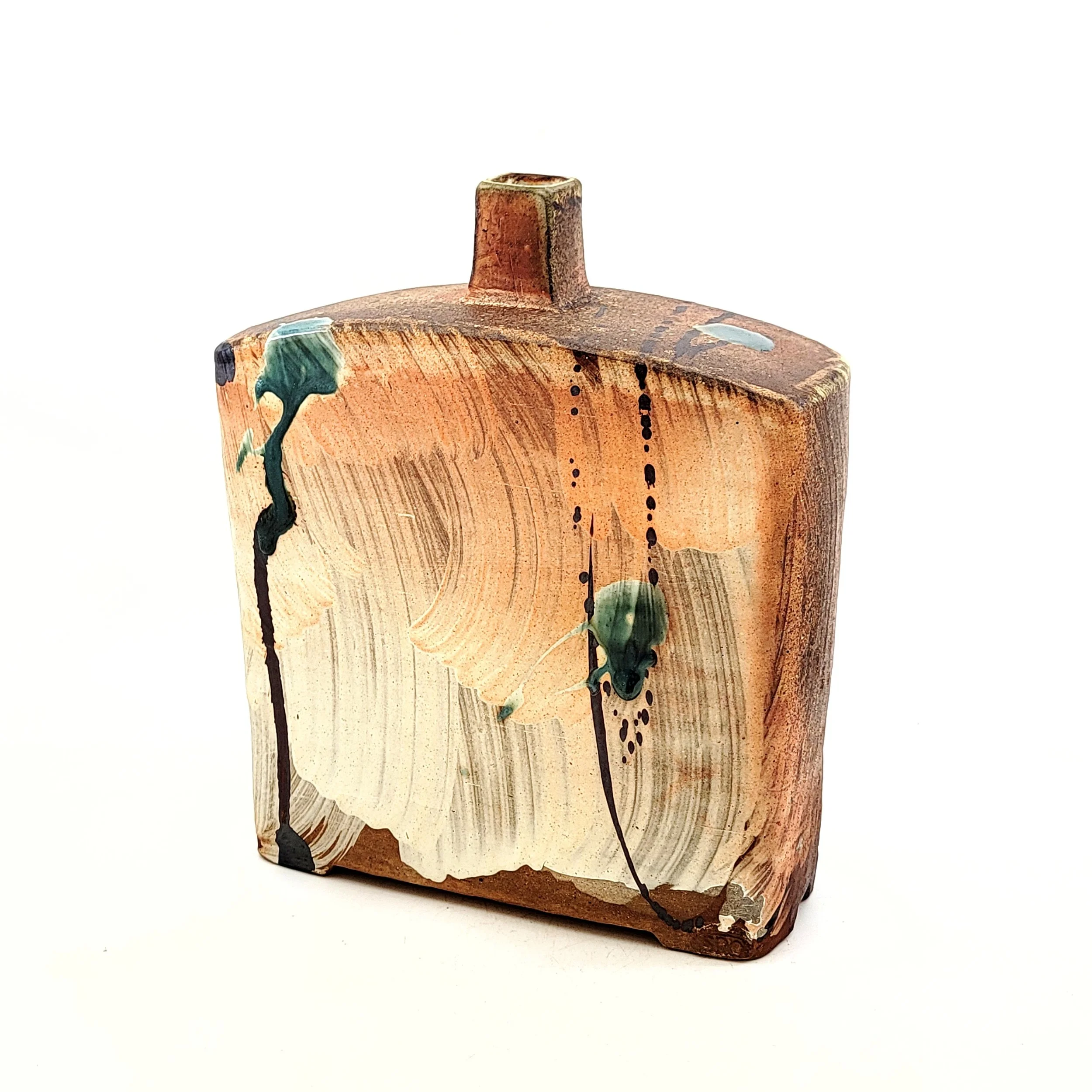 A small rectangular wooden sculpture with painted abstract designs and drips, featuring warm earthy tones and splashes of green, set against a plain white background.