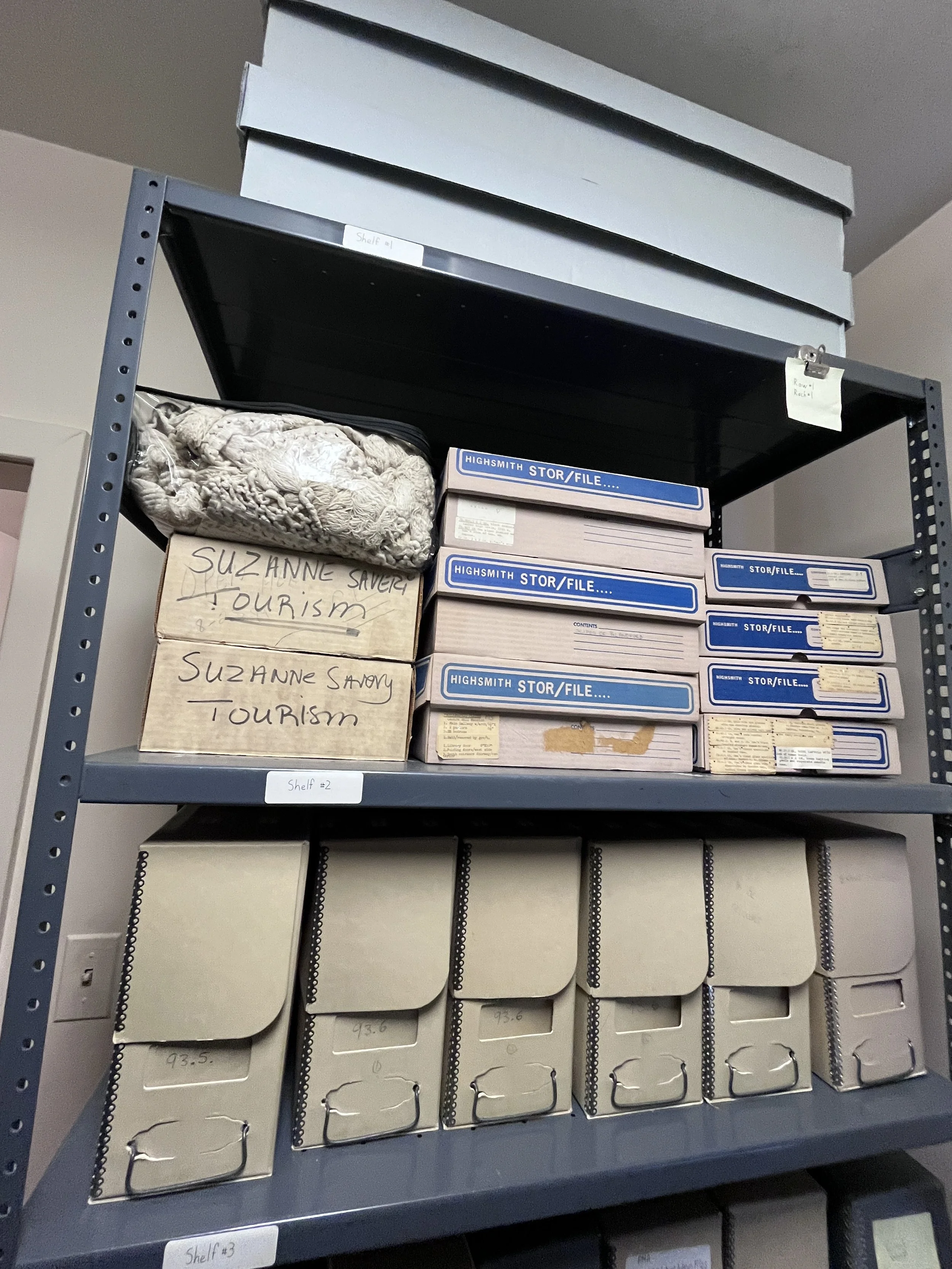 A metal storage shelf holding labeled boxes, folders, and cardboard containers organized on three shelves.