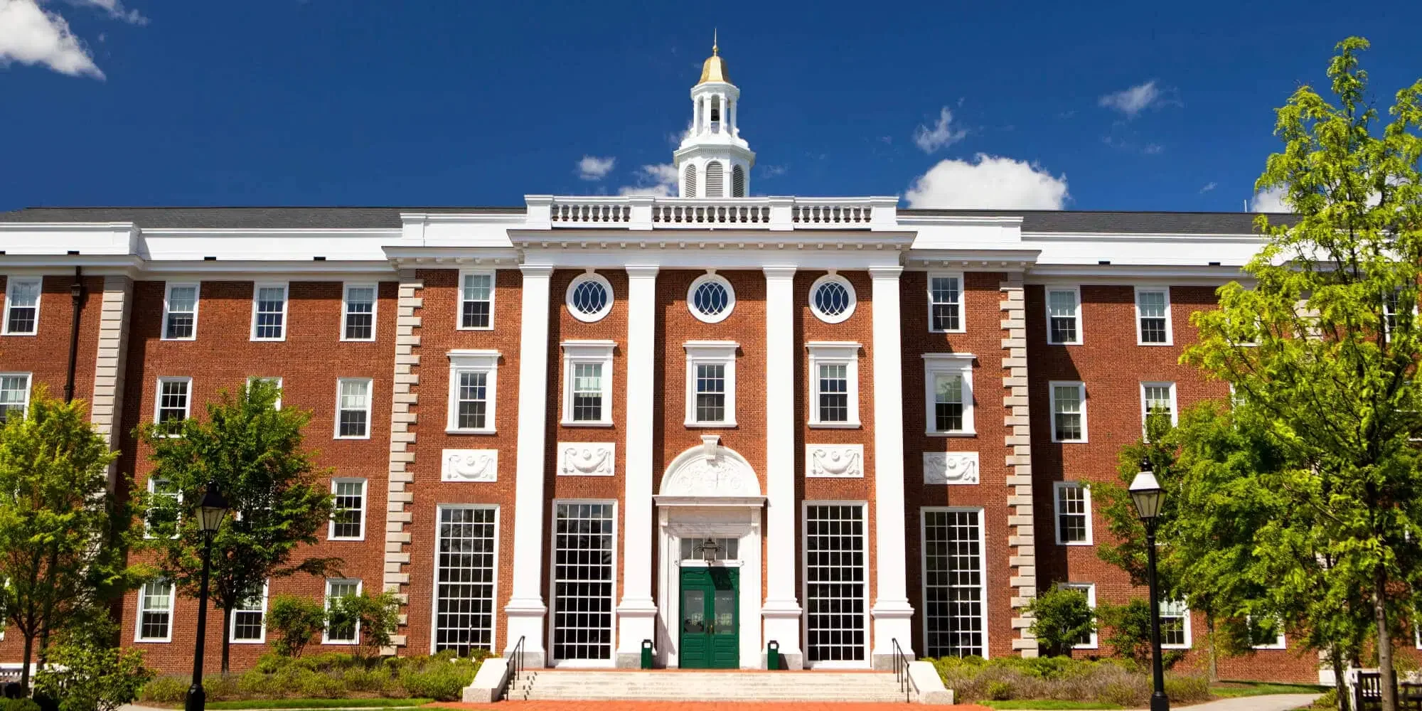 A large brick university building with white architectural details, green door, and a clock tower with a gold spire, surrounded by trees and street lamps, under a partly cloudy blue sky.