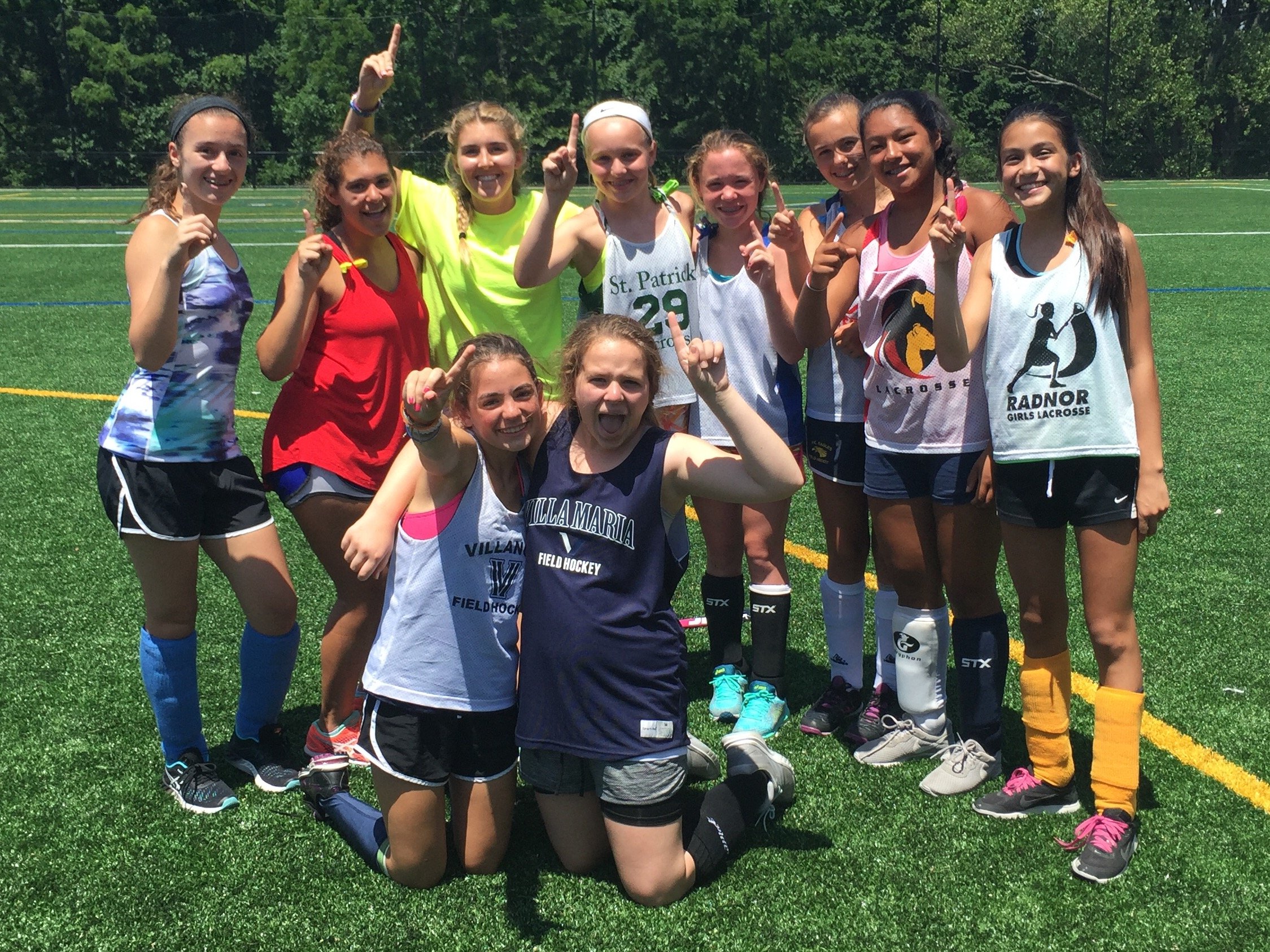 Group of young girls on a sports field, smiling and pointing up, wearing athletic uniforms and track gear.