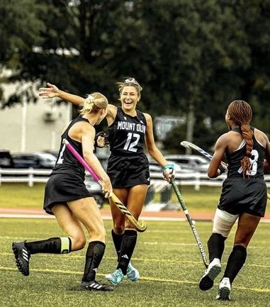 Three female field hockey players in black uniforms celebrating on the field. They are smiling and appear to be happy due to their success on the field.