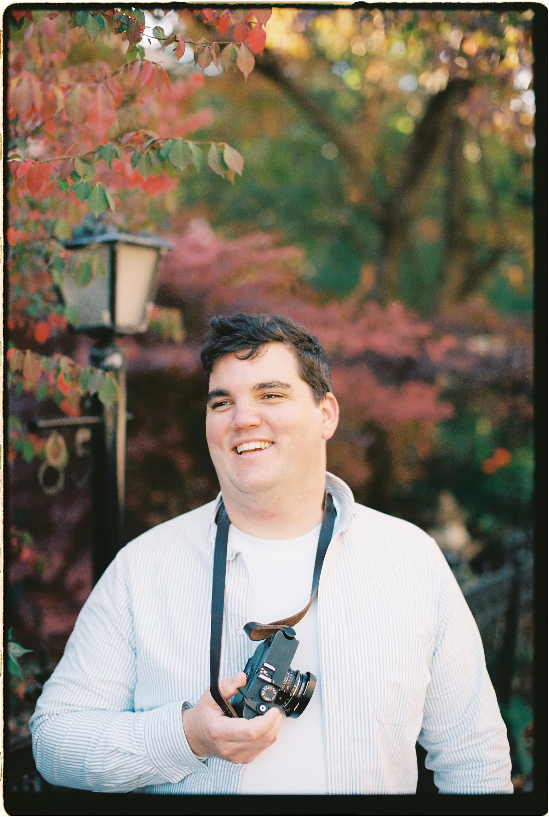 A smiling man holding a camera with a strap around his neck outdoors during fall, with red and orange leaves on trees in the background.