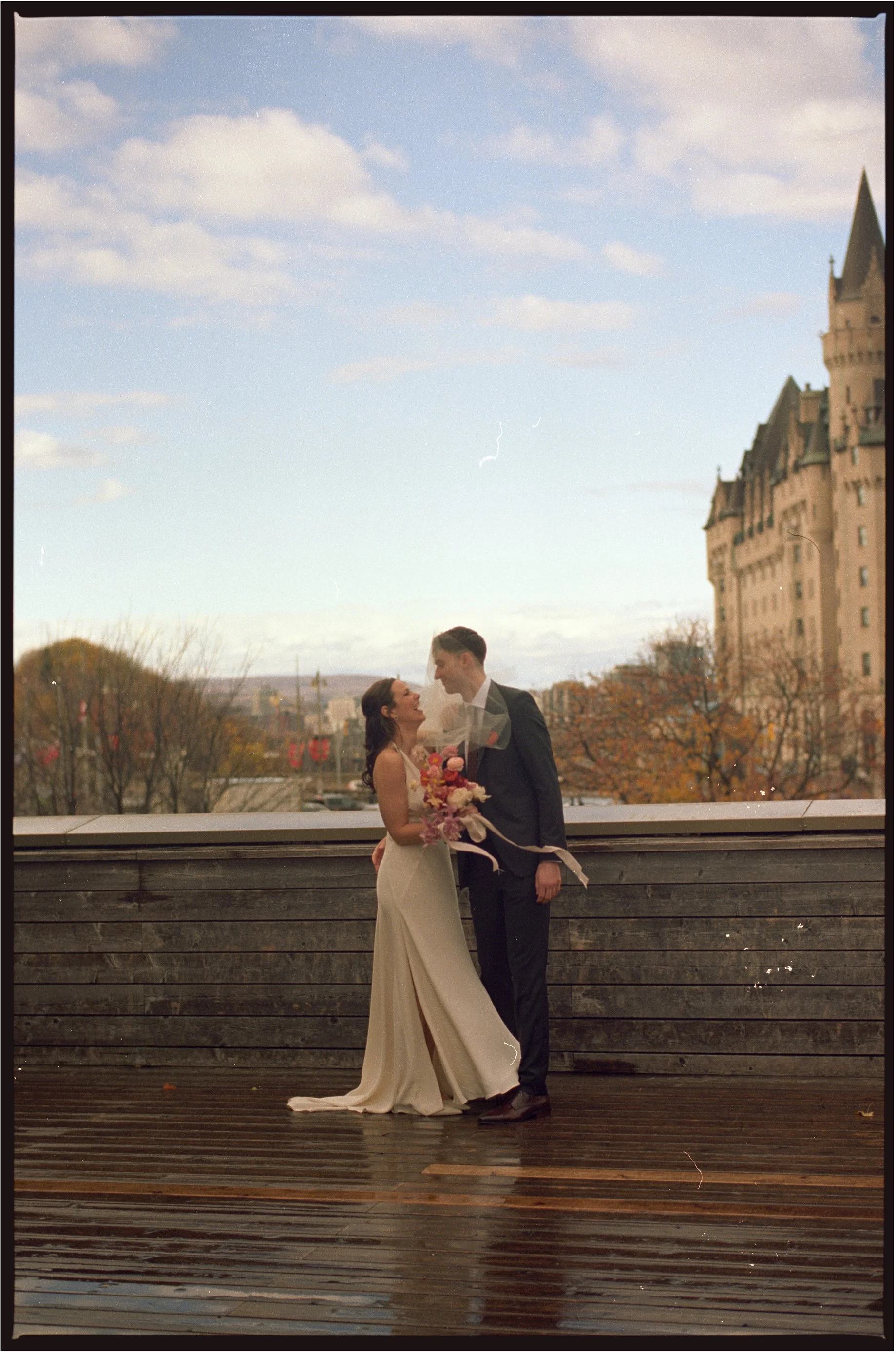 A bride and groom sharing a romantic moment outdoors on a wooden deck, with a castle-like building and trees with autumn foliage in the background.