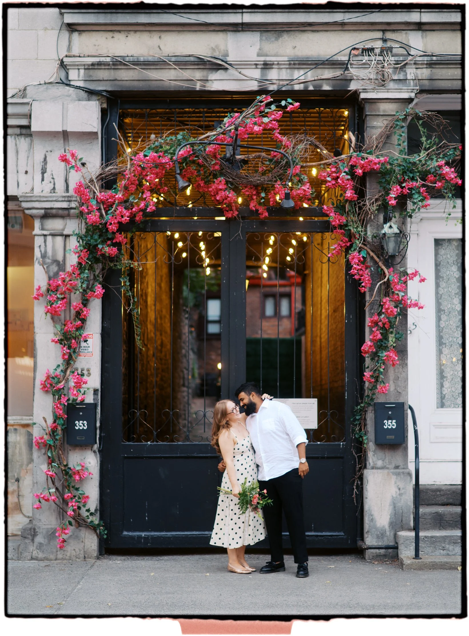 A couple standing close together, smiling and touching foreheads, standing in front of a black gate decorated with pink flowers and fairy lights. The woman is holding a bouquet of flowers, and the man is dressed in a white shirt and black pants.