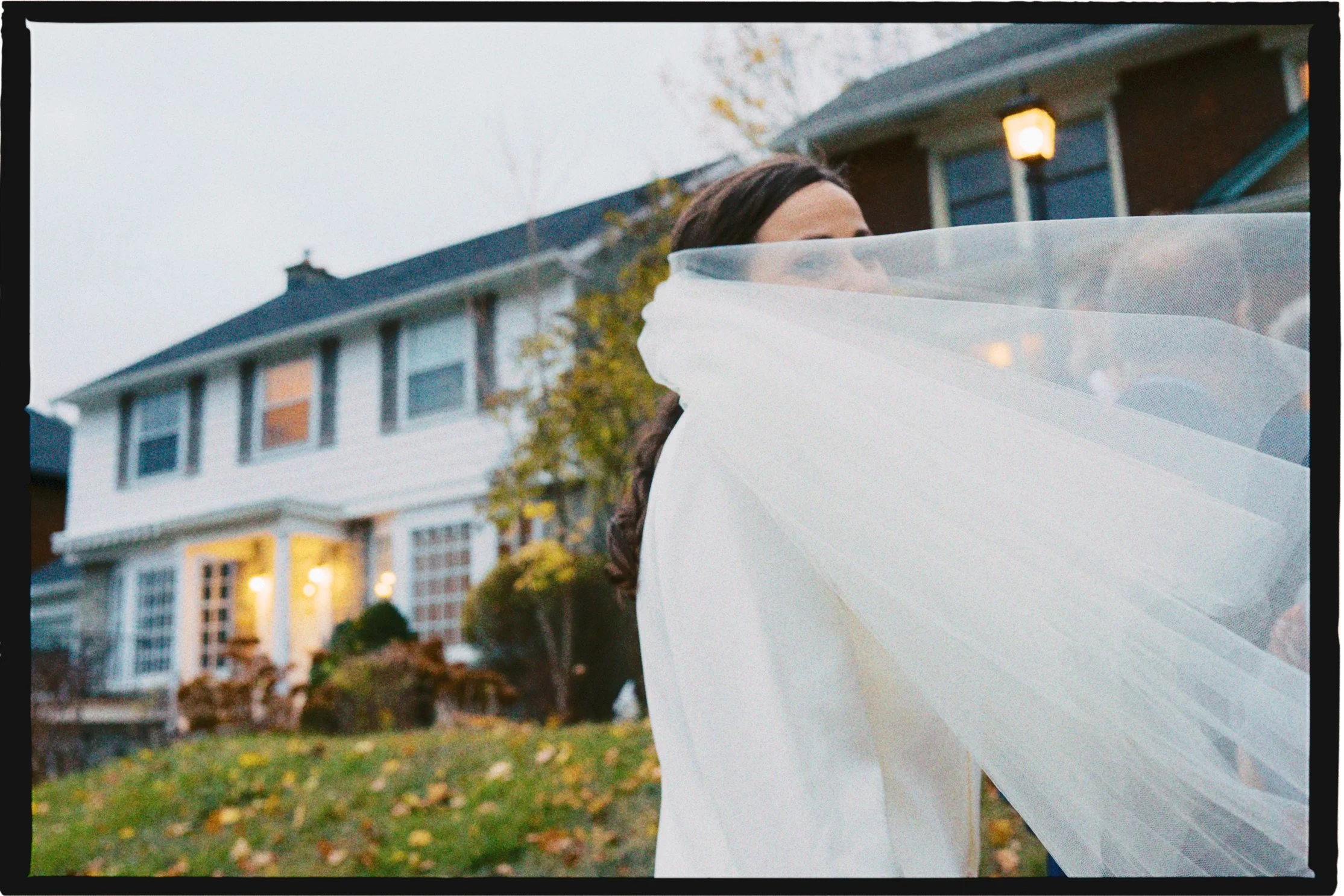 A woman with brown hair in a white wedding dress standing outside in front of a large house with lights on and autumn leaves on the ground, with part of her veil blowing in the wind.