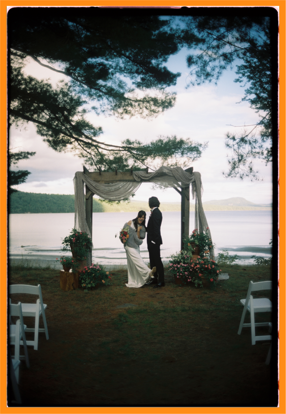 A couple getting married outdoors under a wooden arch decorated with draped fabric and flowers, near a lake with trees and mountains in the background.