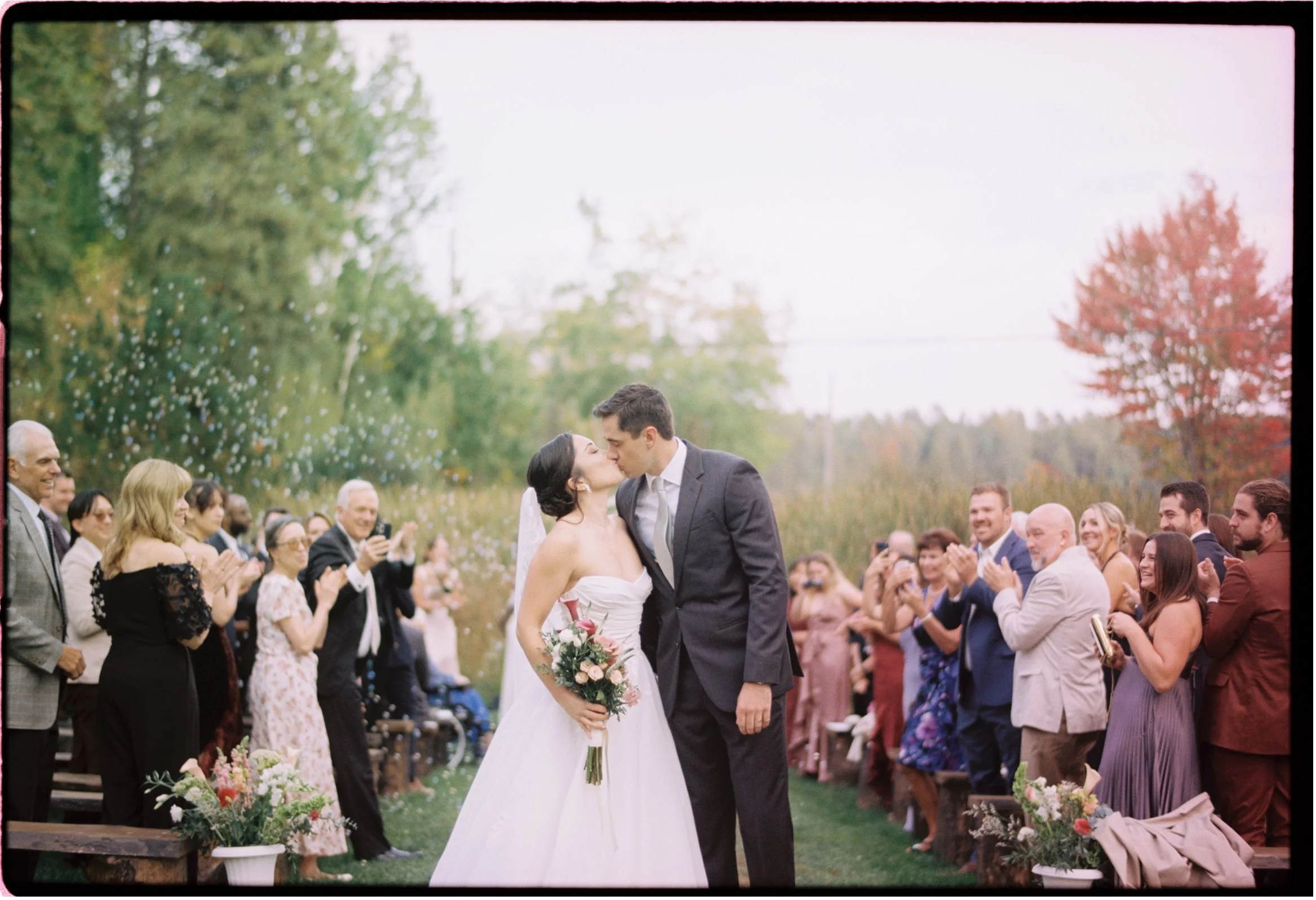 Ottawa Wedding Photo, film wedding, Kodak wedding, 35mm wedding. A bride and groom kiss as they leave their outdoor wedding ceremony, surrounded by friends and family clapping and celebrating in a natural setting with trees and colorful foliage.
