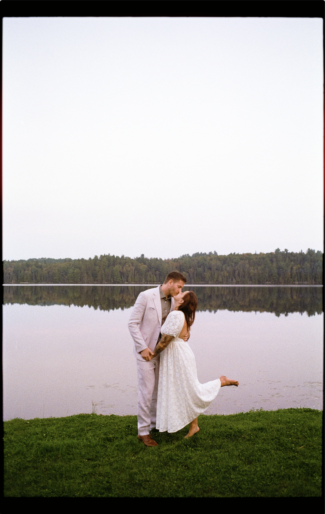 A man and woman kiss by a lake. The woman lifts her leg as they hold hands on a grassy area.