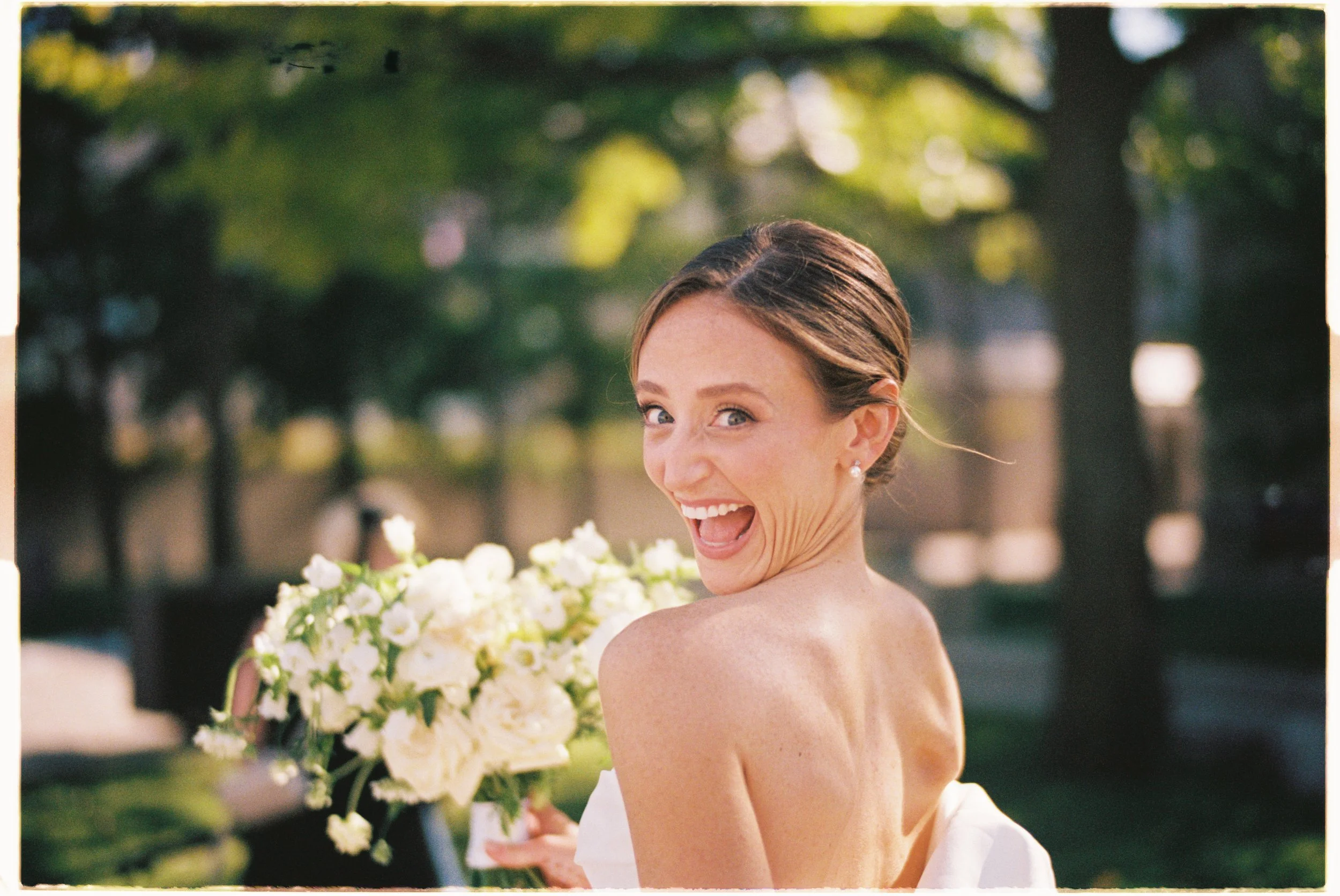 A woman smiling and looking back over her shoulder outdoors holding a bouquet of white flowers, with trees and blurred background.
