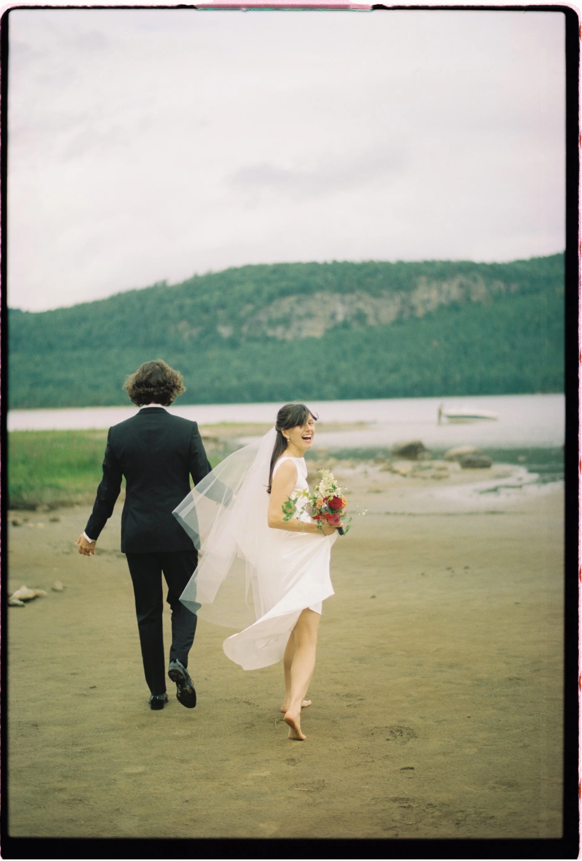 A wedding photo of a bride and groom walking on a beach with a river, rocks, and green hills in the background.