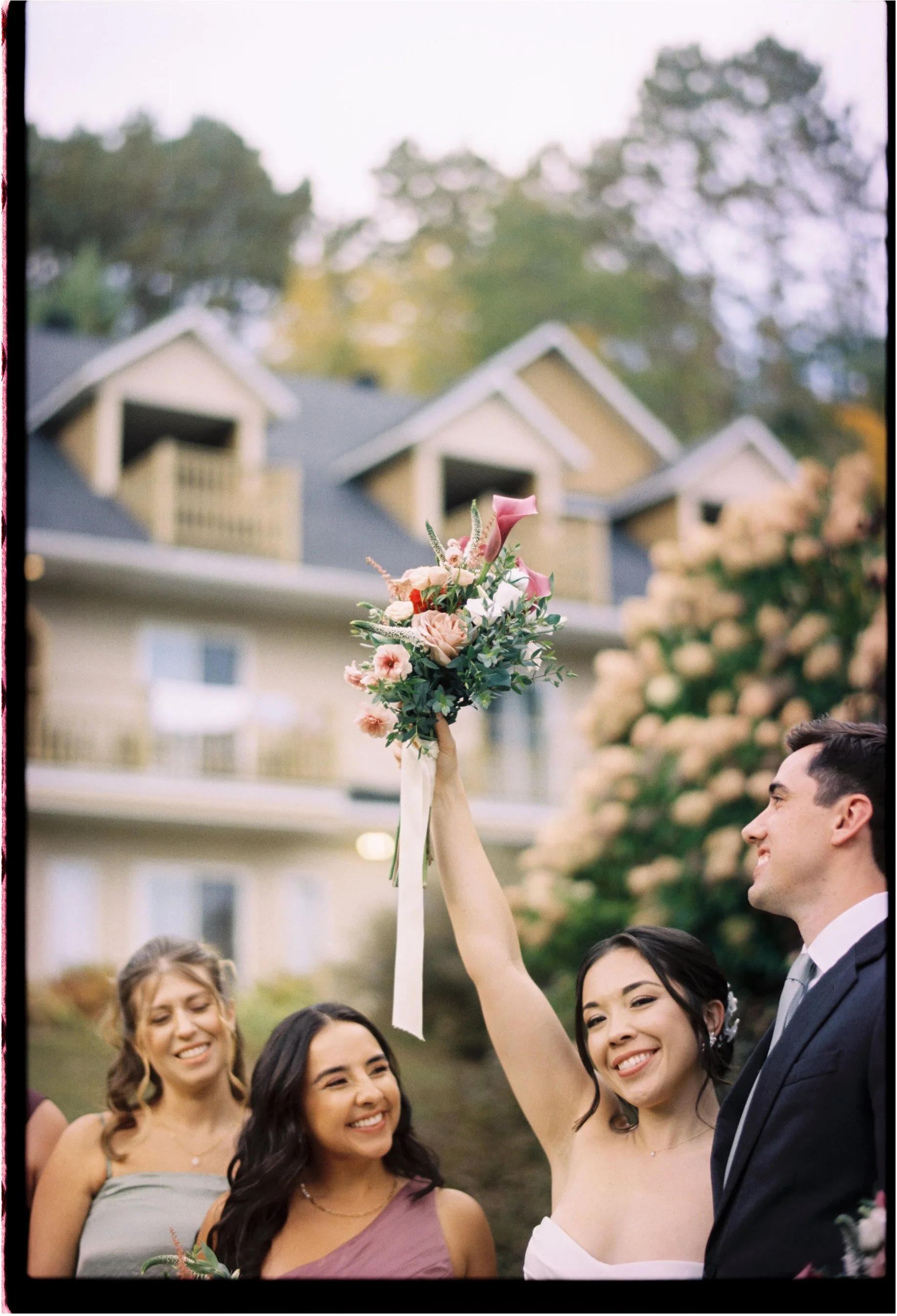 A bride in a white dress holds up a bouquet of pink and white flowers during a wedding ceremony outside, with a smiling groom in a dark suit beside her, turned toward her. Two bridesmaids are in the background, standing in front of a building with mu