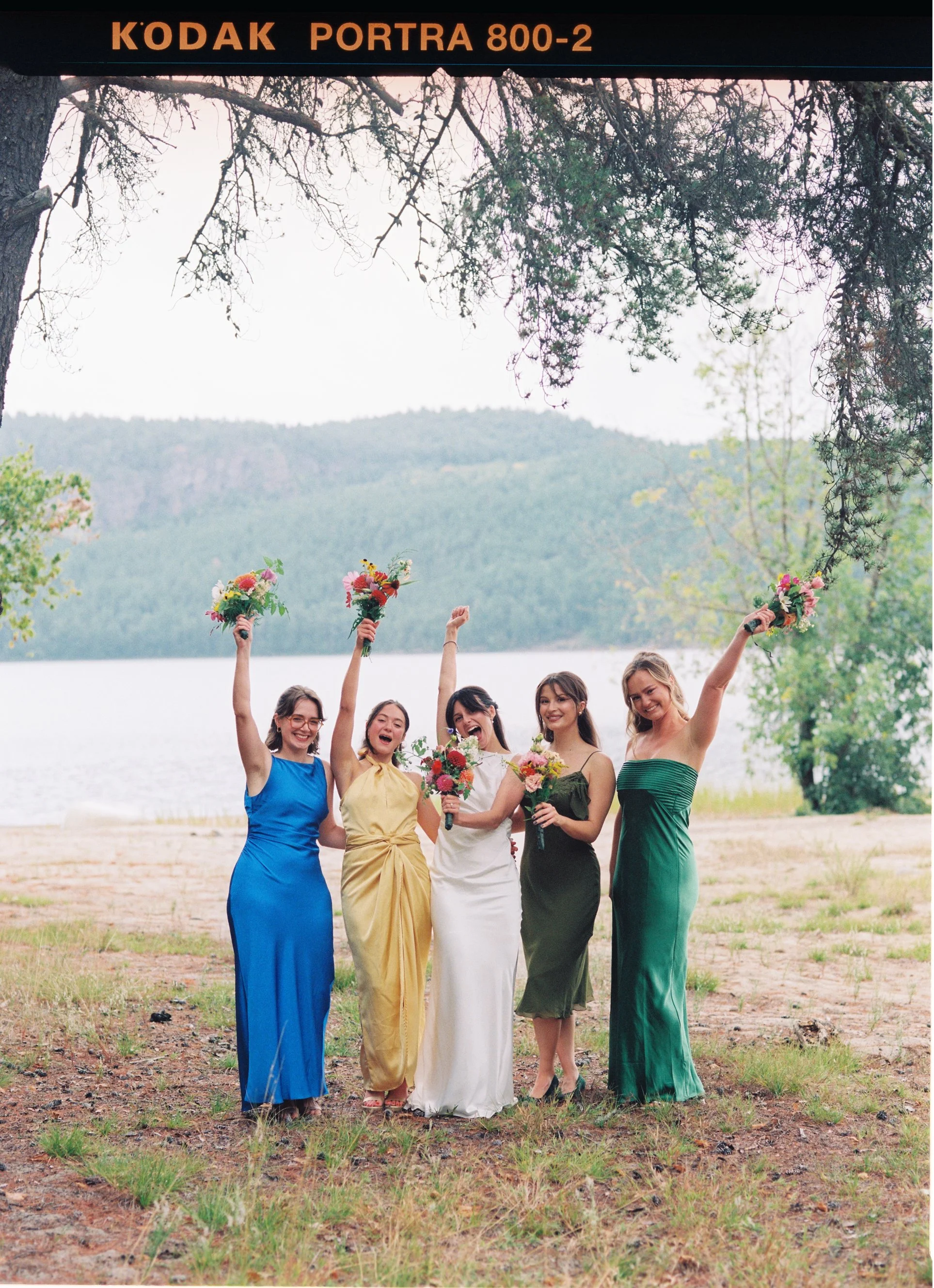 A group of five women in colorful dresses holding bouquets of flowers, celebrating outdoors near a lake with trees and mountains in the background.