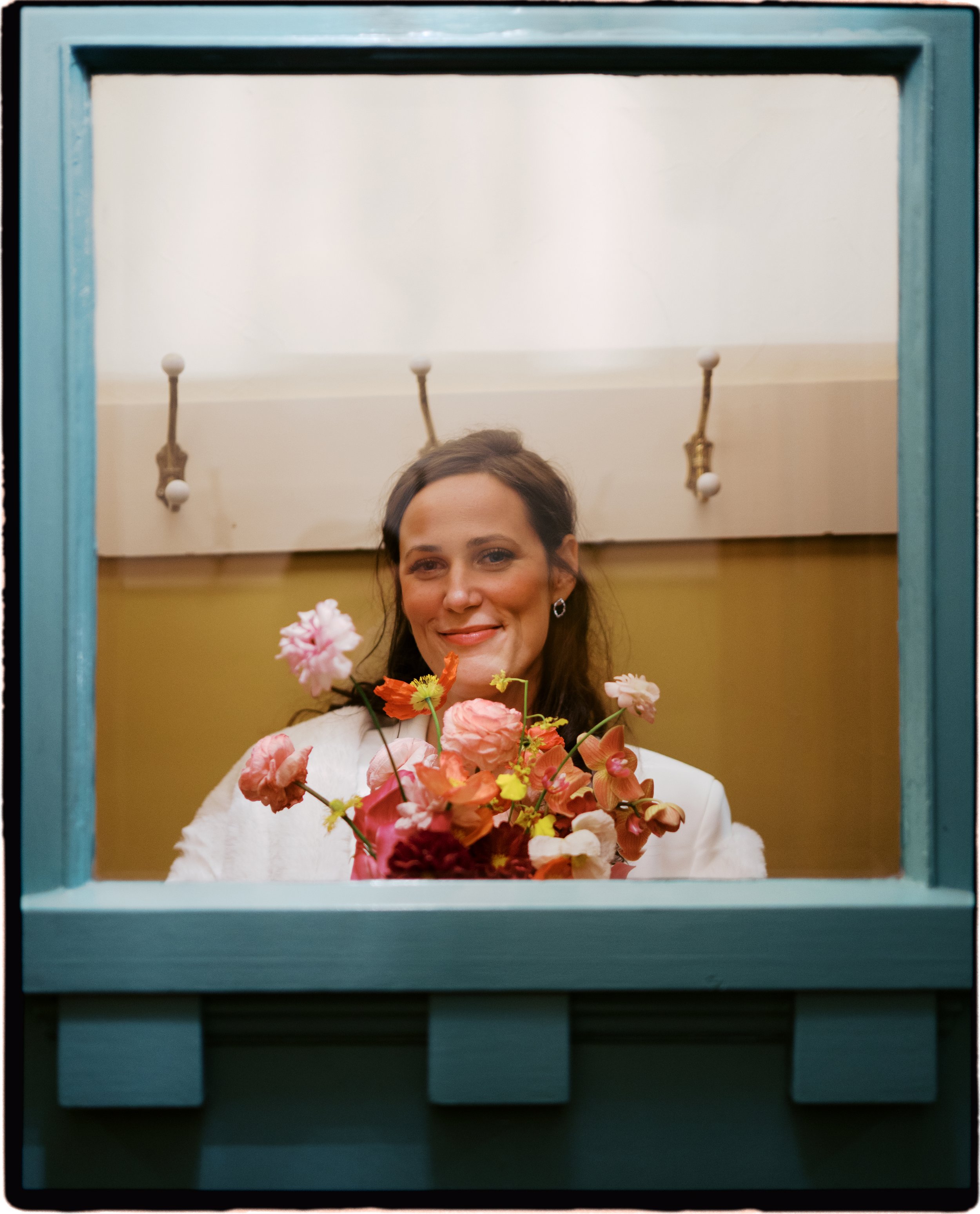 A woman smiling in a mirror holds a bouquet of pink and orange flowers. She has dark hair, is wearing earrings, and a white outfit. The mirror has a turquoise frame, and there are two wall-mounted light fixtures in the background.