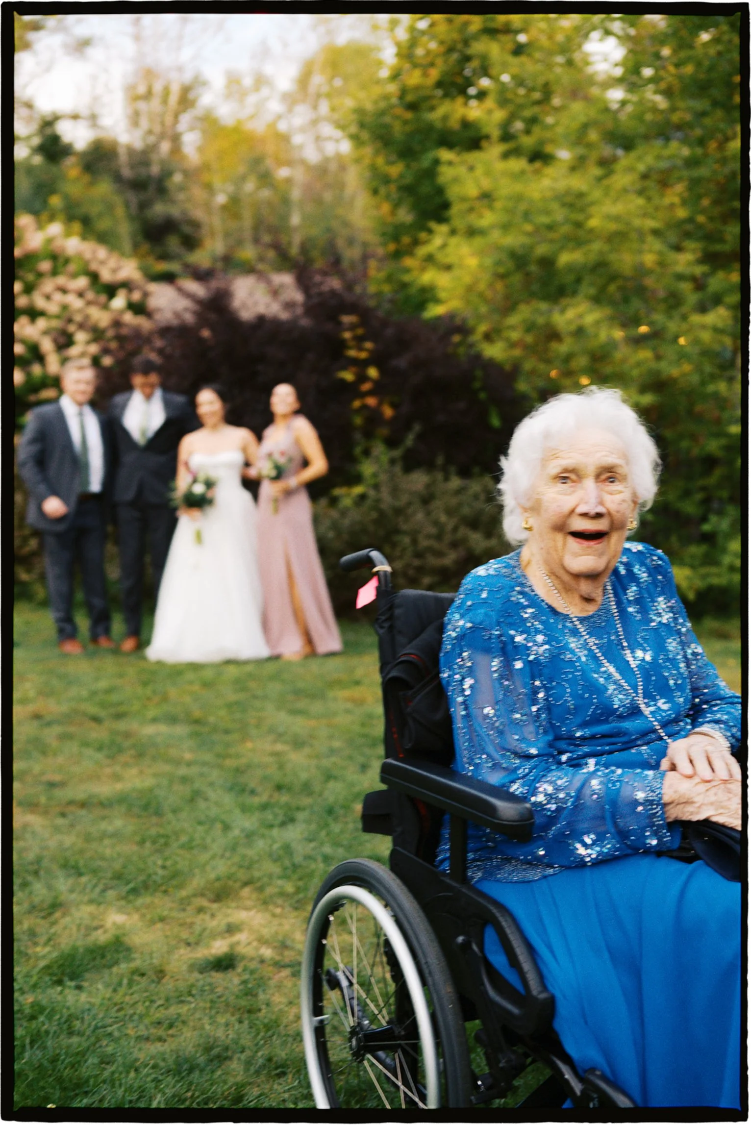 An elderly woman in a wheelchair smiling in the foreground, with a wedding party blurred in the background outdoors among trees.