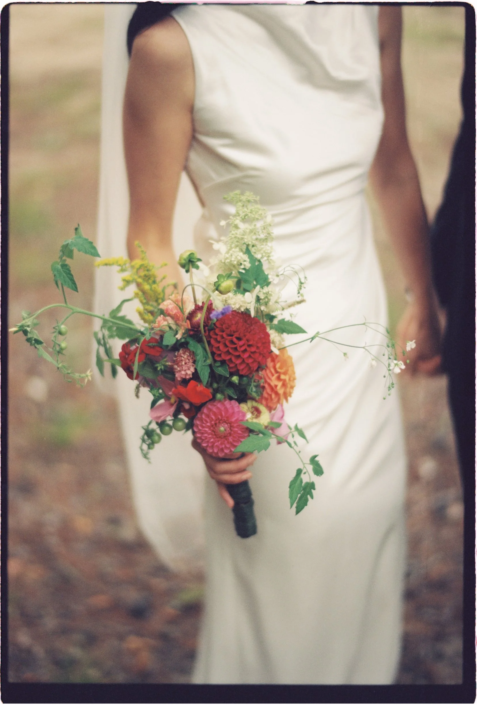 A bride in a sleeveless white wedding dress holding a colorful bouquet of flowers, standing outdoors on a natural ground surface.