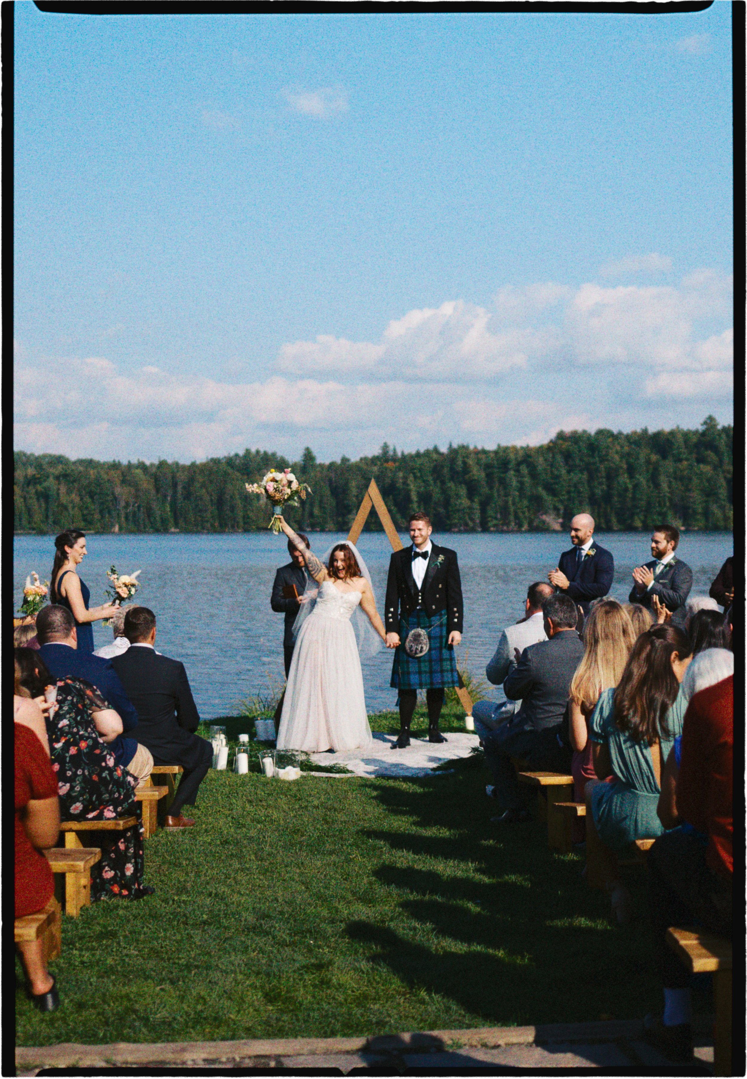A wedding ceremony taking place outdoors by a lake, with the bride and groom standing together and holding hands. The bride is holding a bouquet in the air, and the groom is wearing a kilt. Guests are seated on benches, watching and clapping. The bac