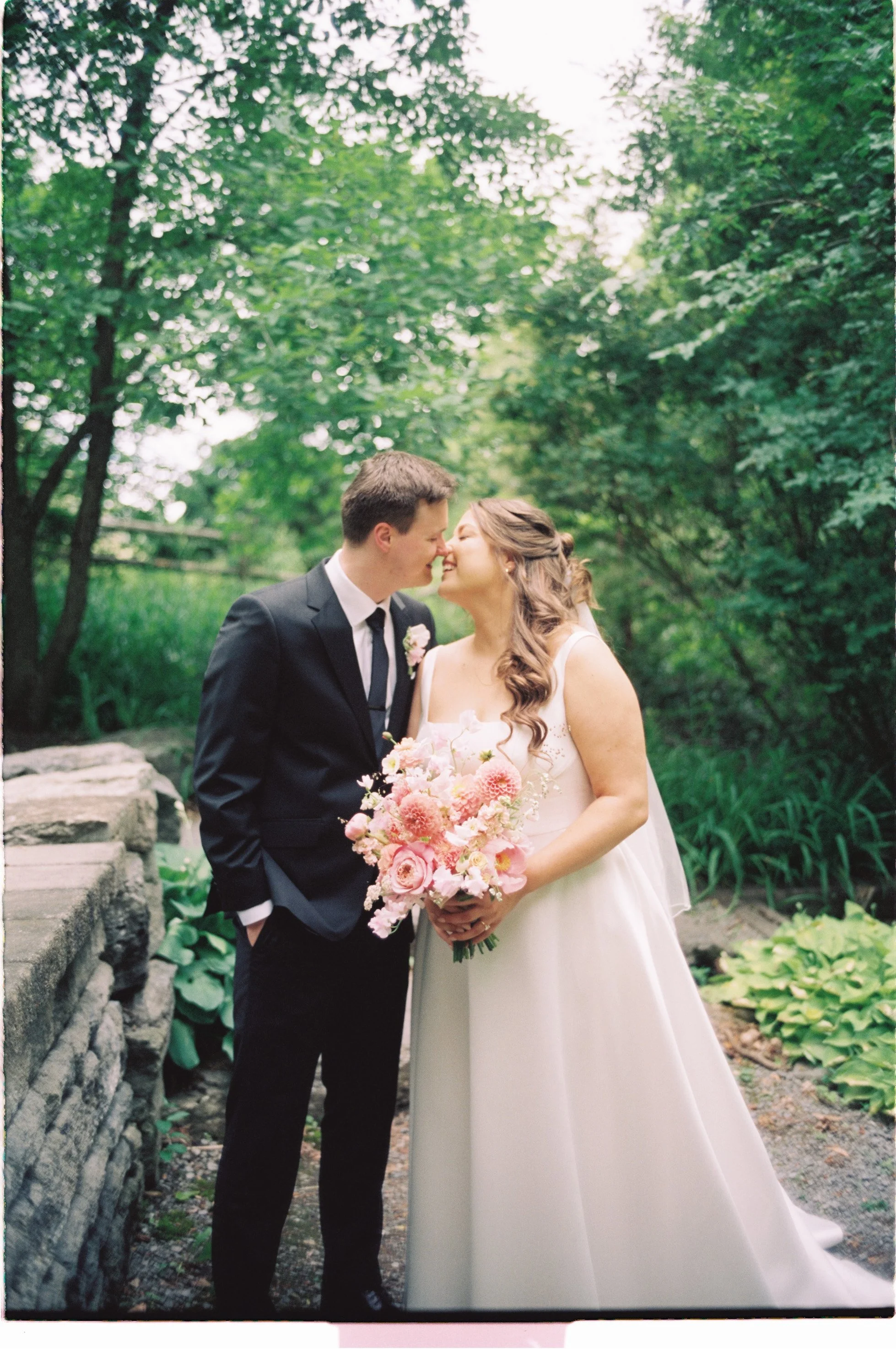A bride and groom on their wedding day, standing outdoors among green trees, leaning close, holding a bouquet of pink flowers, with the bride in a white gown and the groom in a black suit, smiling gently at each other.