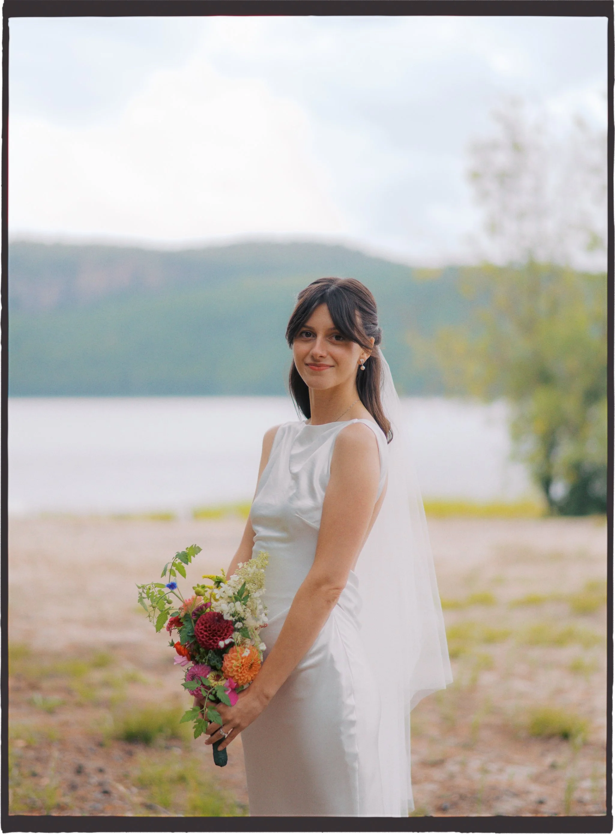 A bride holding a colorful bouquet of flowers outdoors near a lake with mountains in the background.