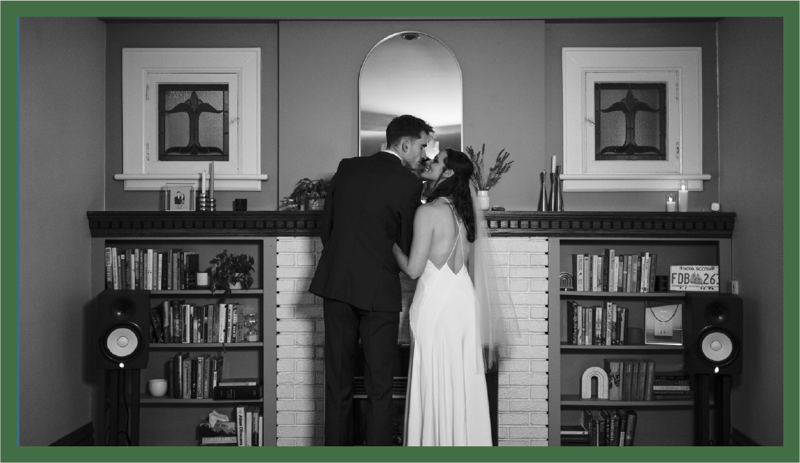 A couple dressed in wedding attire is kissing in front of a fireplace with bookshelves, candles, and decorative items in the background.