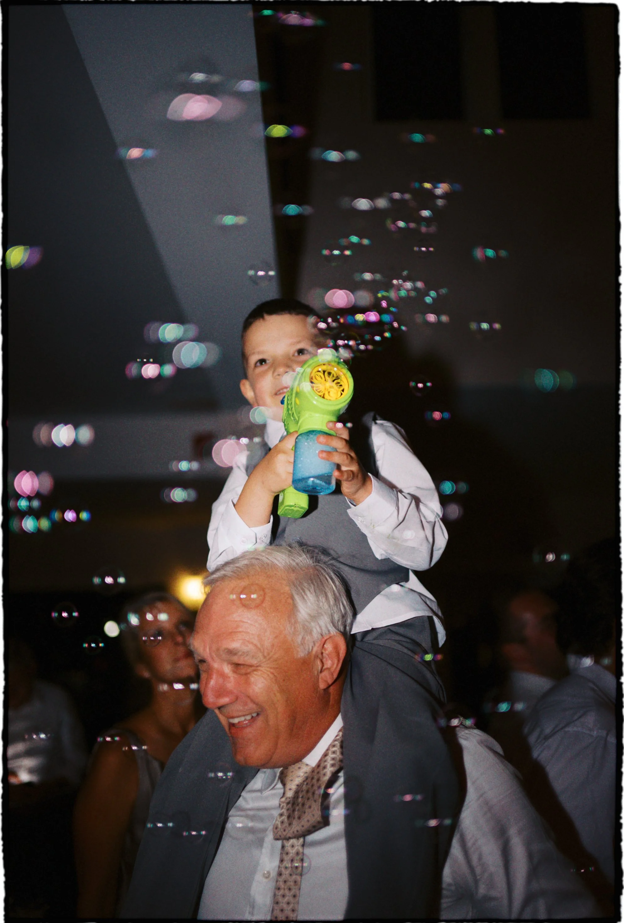 A young boy sitting on an older man's shoulders, playing with a bubble gun, with bubbles floating around in a dimly lit room.Ottawa Wedding Photo, film wedding, Kodak wedding, 35mm wedding