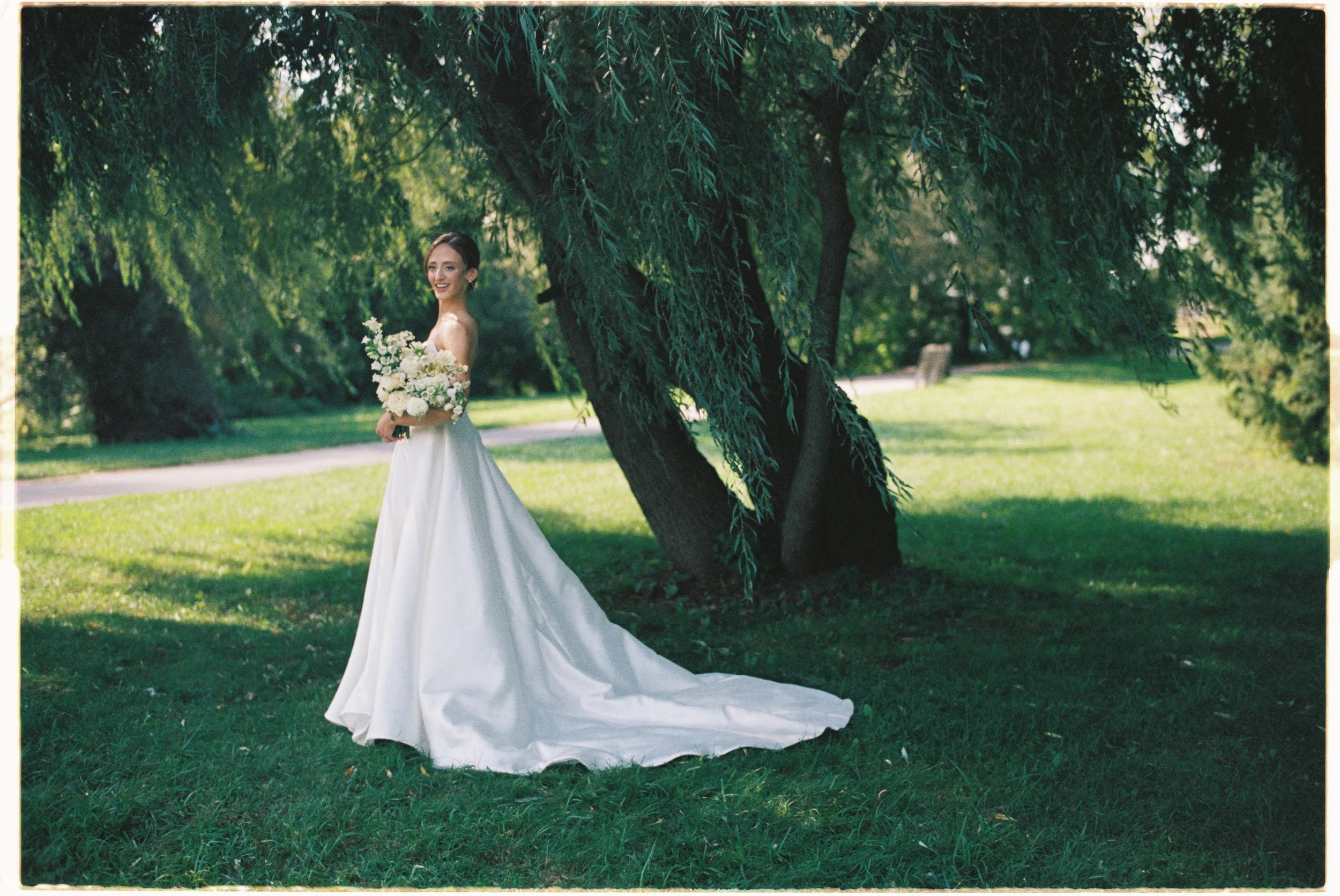 A bride standing on grass in a park, holding a bouquet of white flowers, wearing a white wedding dress with a long train, under a large tree with lush green leaves.
