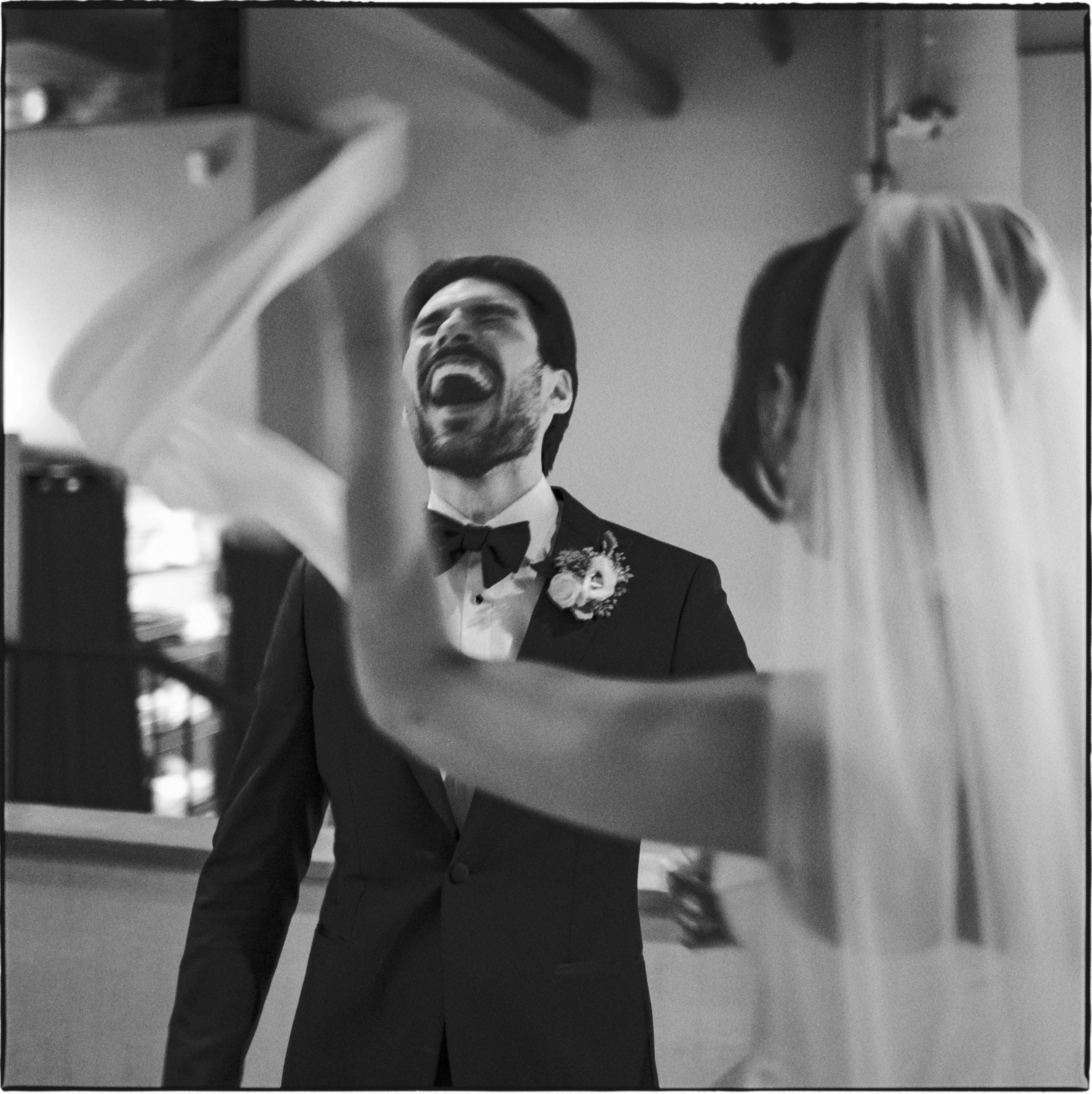 A groom in a tuxedo laughing while a bride in a wedding dress, with her back to the camera, is dancing or celebrating at a wedding reception.