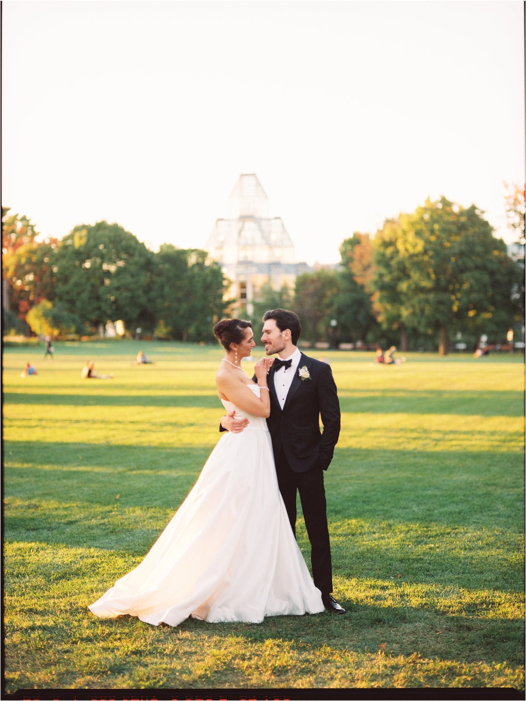 Ottawa Wedding Photo, film wedding, Kodak wedding, 35mm wedding A bride and groom in wedding attire standing on a grassy field, smiling and embracing during sunset, with trees and a modern building in the background.