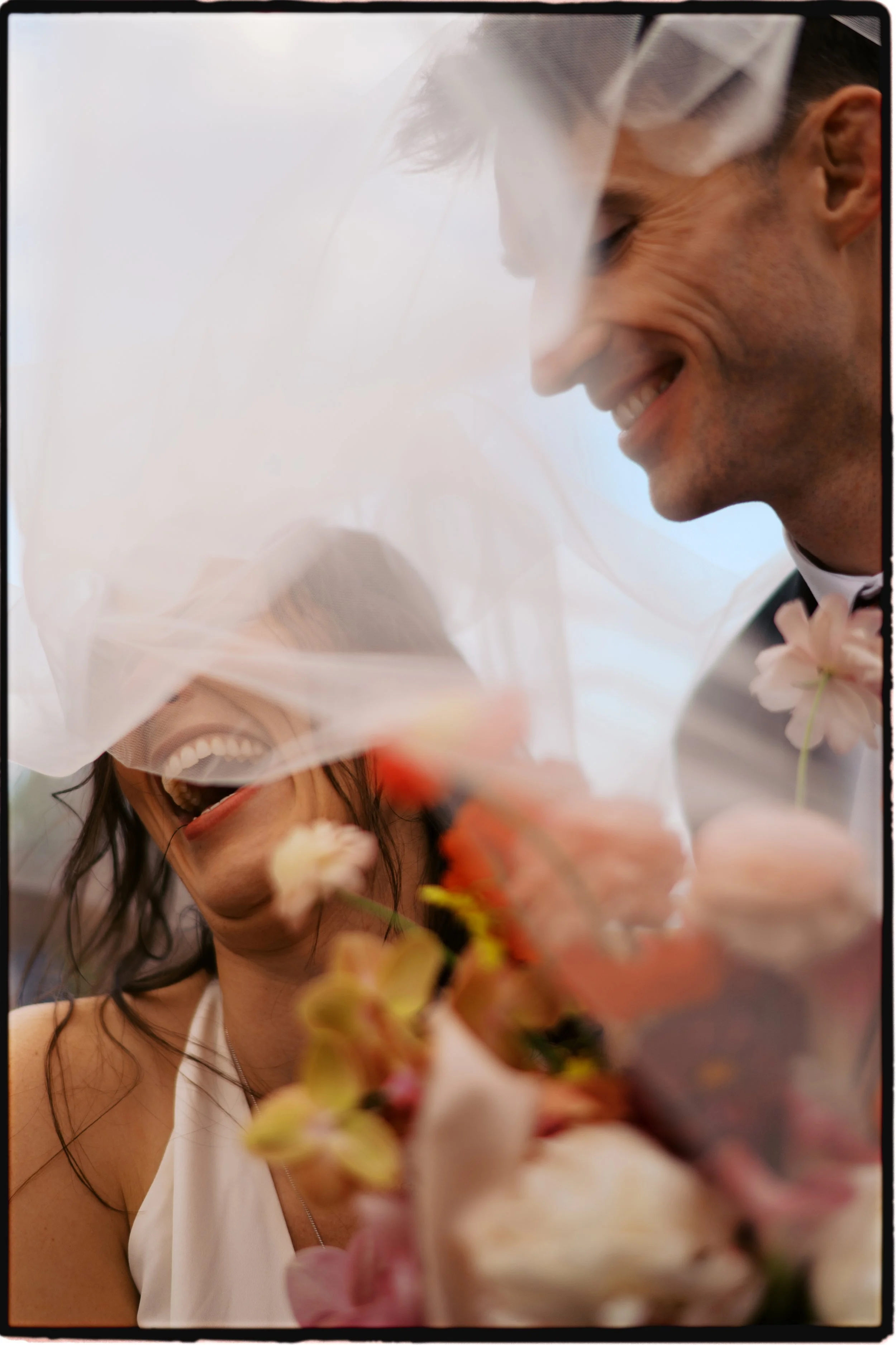 A bride and groom smiling and laughing under a veil, celebrating their wedding with colorful flowers in the foreground.Ottawa Wedding Photo, film wedding, Kodak wedding, 35mm wedding
