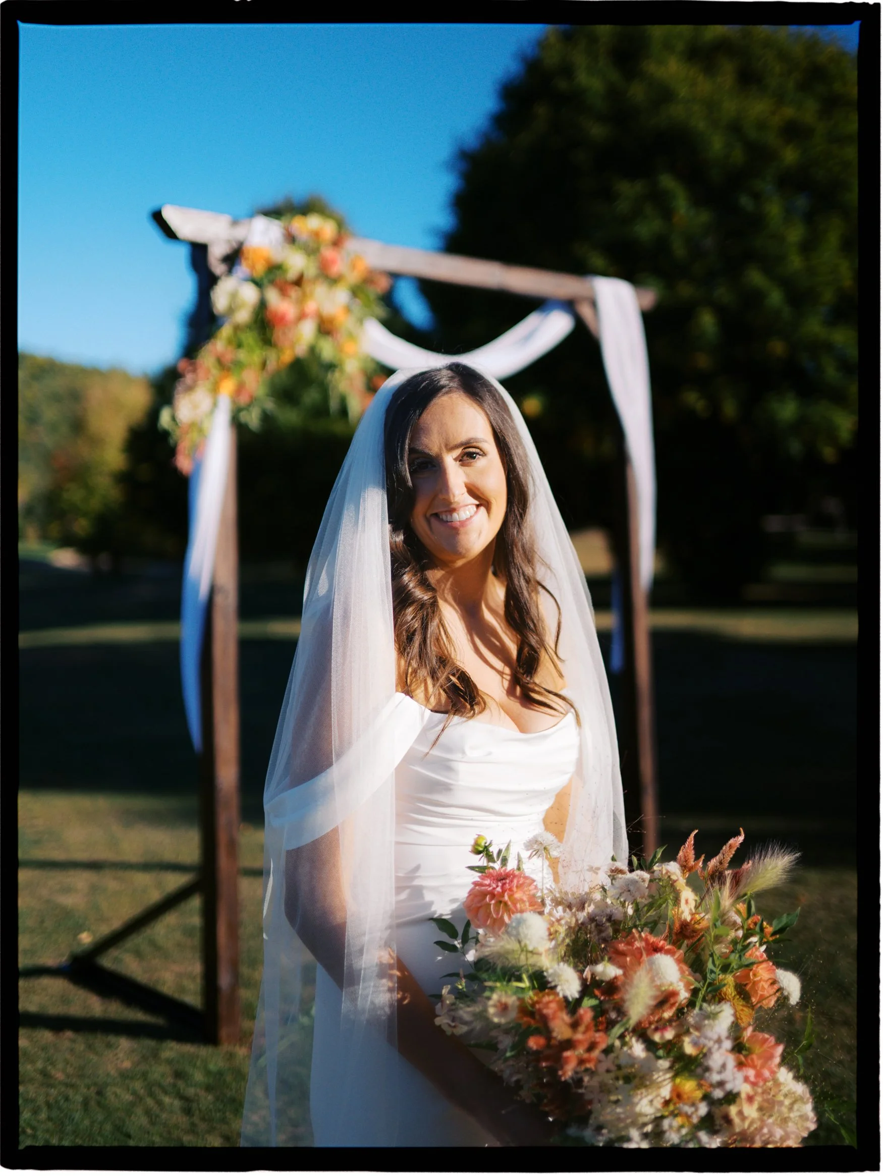 A smiling bride in a white wedding dress with a veil, holding a bouquet of flowers, standing outdoors under a wooden wedding arch decorated with flowers and draped fabric.