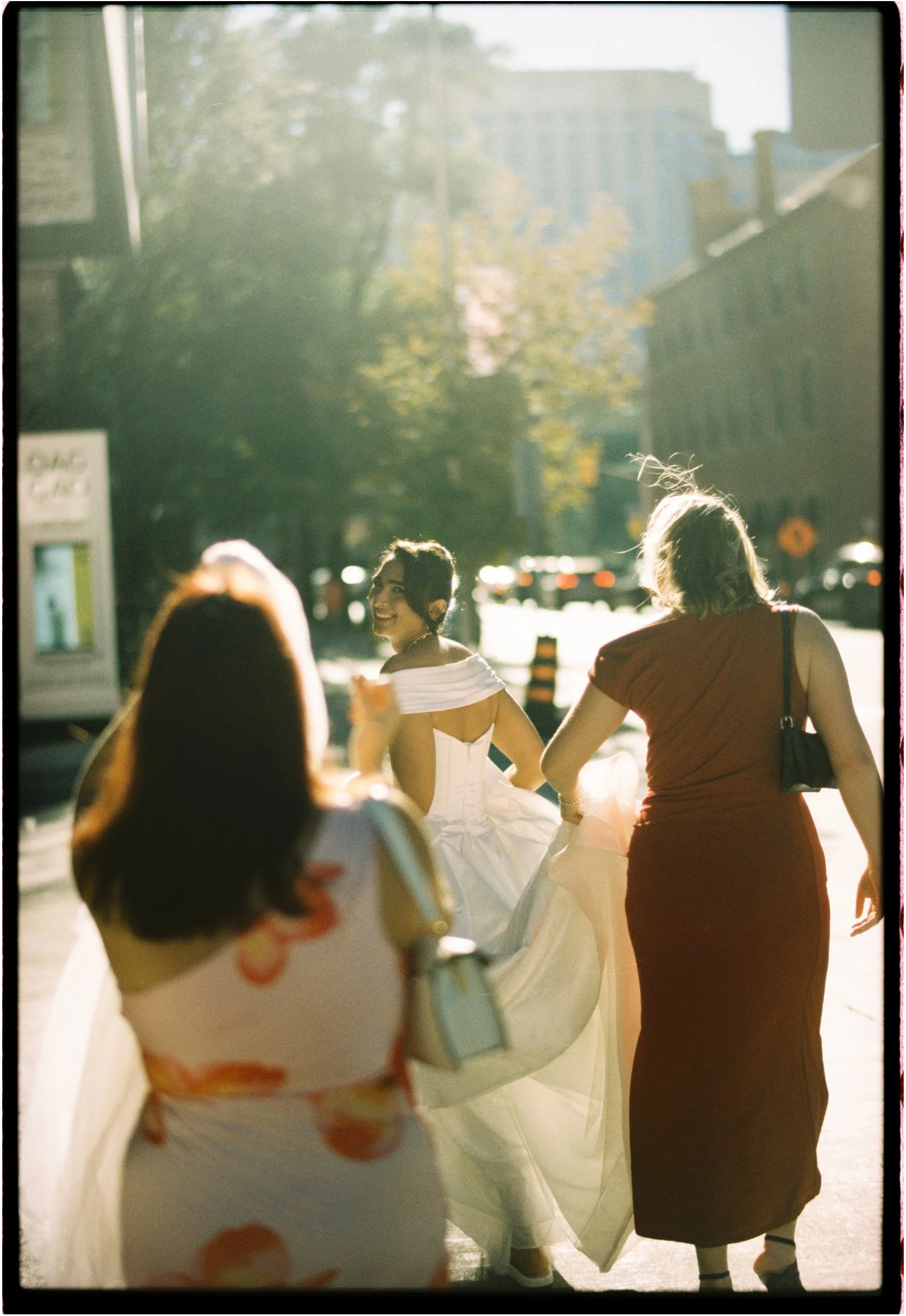 A woman in a white wedding dress walking down the street with two women, one in a red dress and another in a floral dress, on a sunny day.