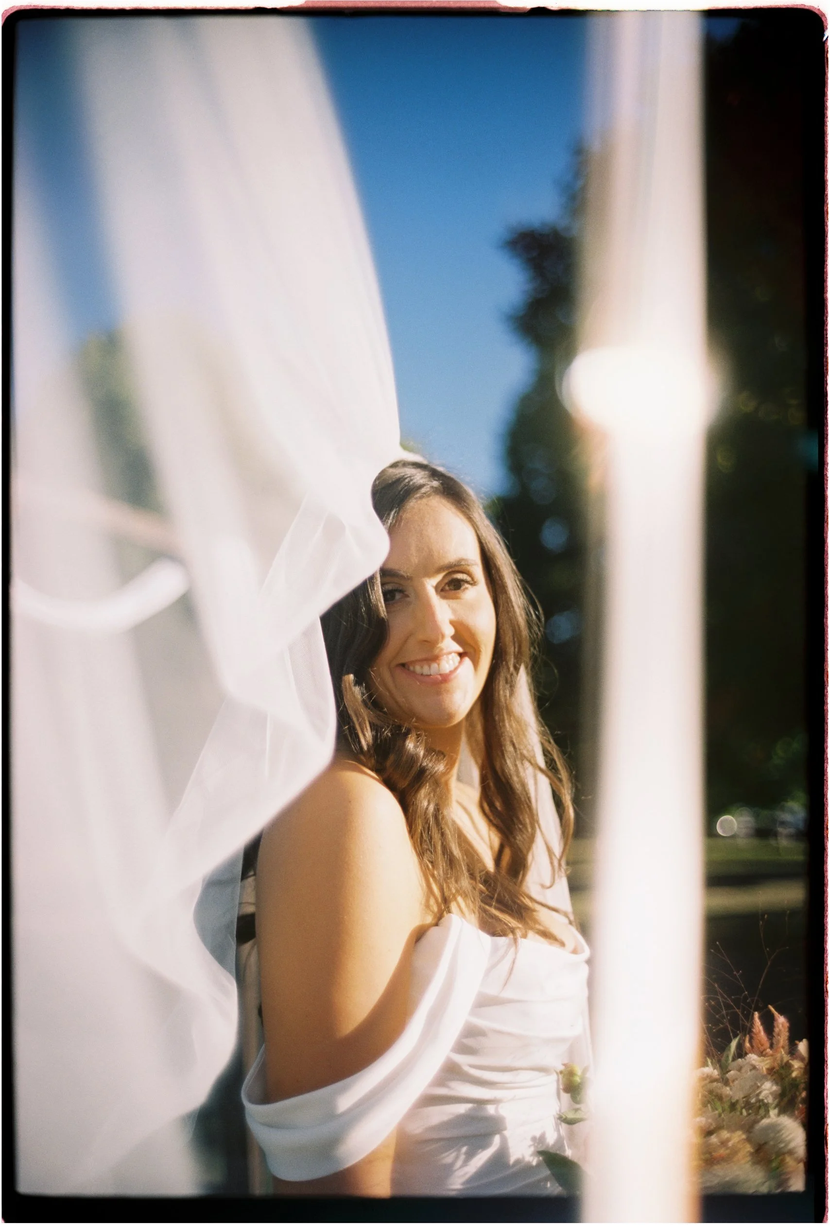 Portrait of a smiling woman in a white dress, with long wavy hair, outdoors on a sunny day, with blurred background of trees and sky.