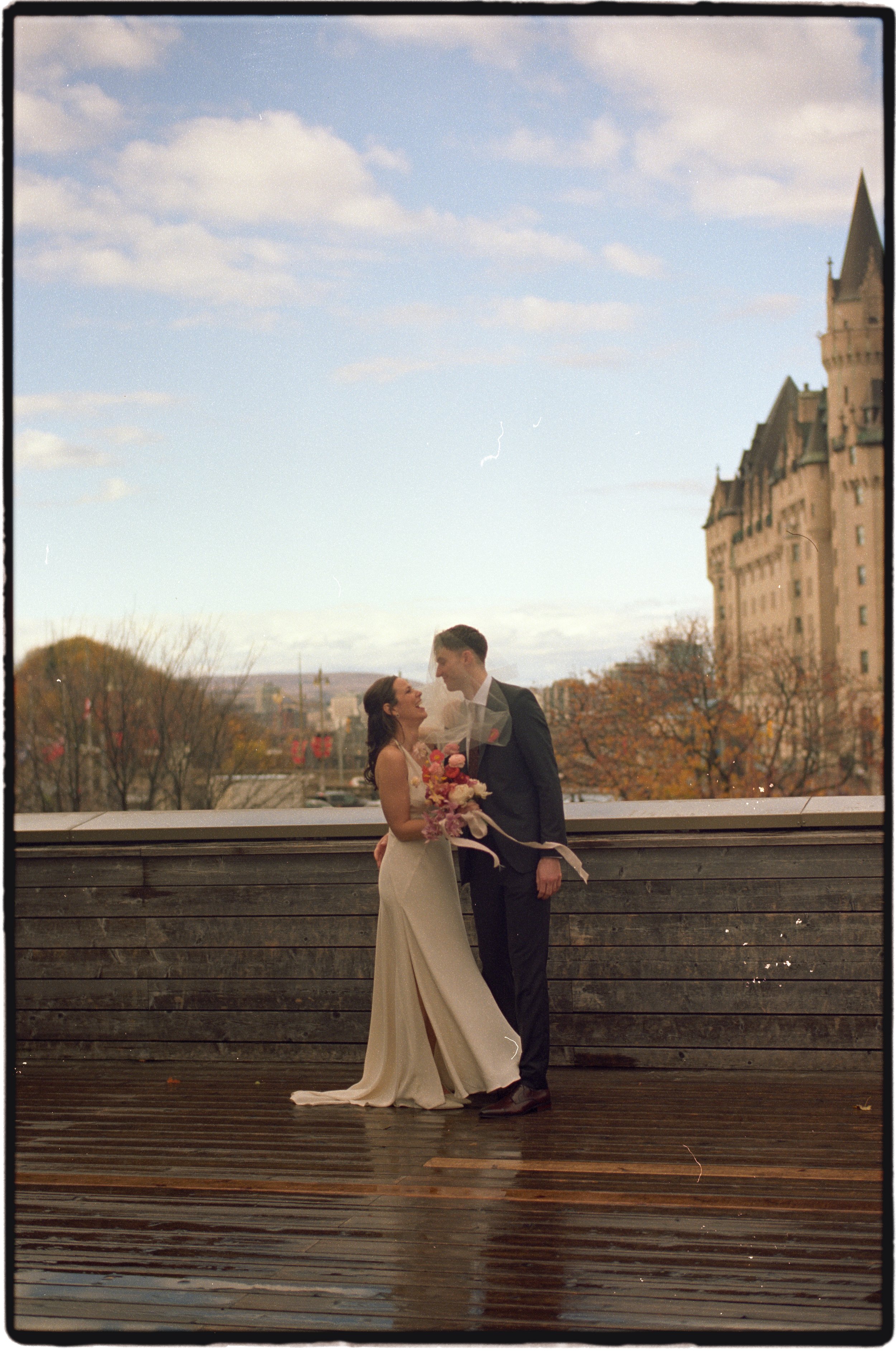Ottawa Wedding Photo, film wedding, Kodak wedding, 35mm wedding bride in a white wedding gown and a groom in a dark suit hold and gaze at each other while standing on a wet wooden deck outdoors, with a cityscape and cloudy sky in the background.