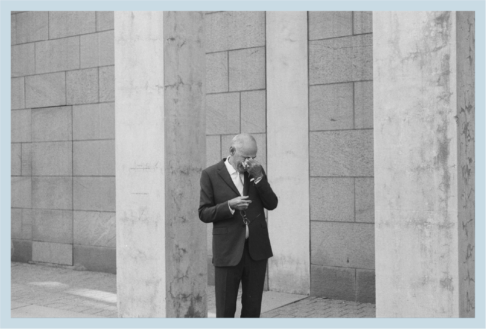 A man in a suit standing against a concrete wall, looking at a smartphone and holding a pair of glasses.