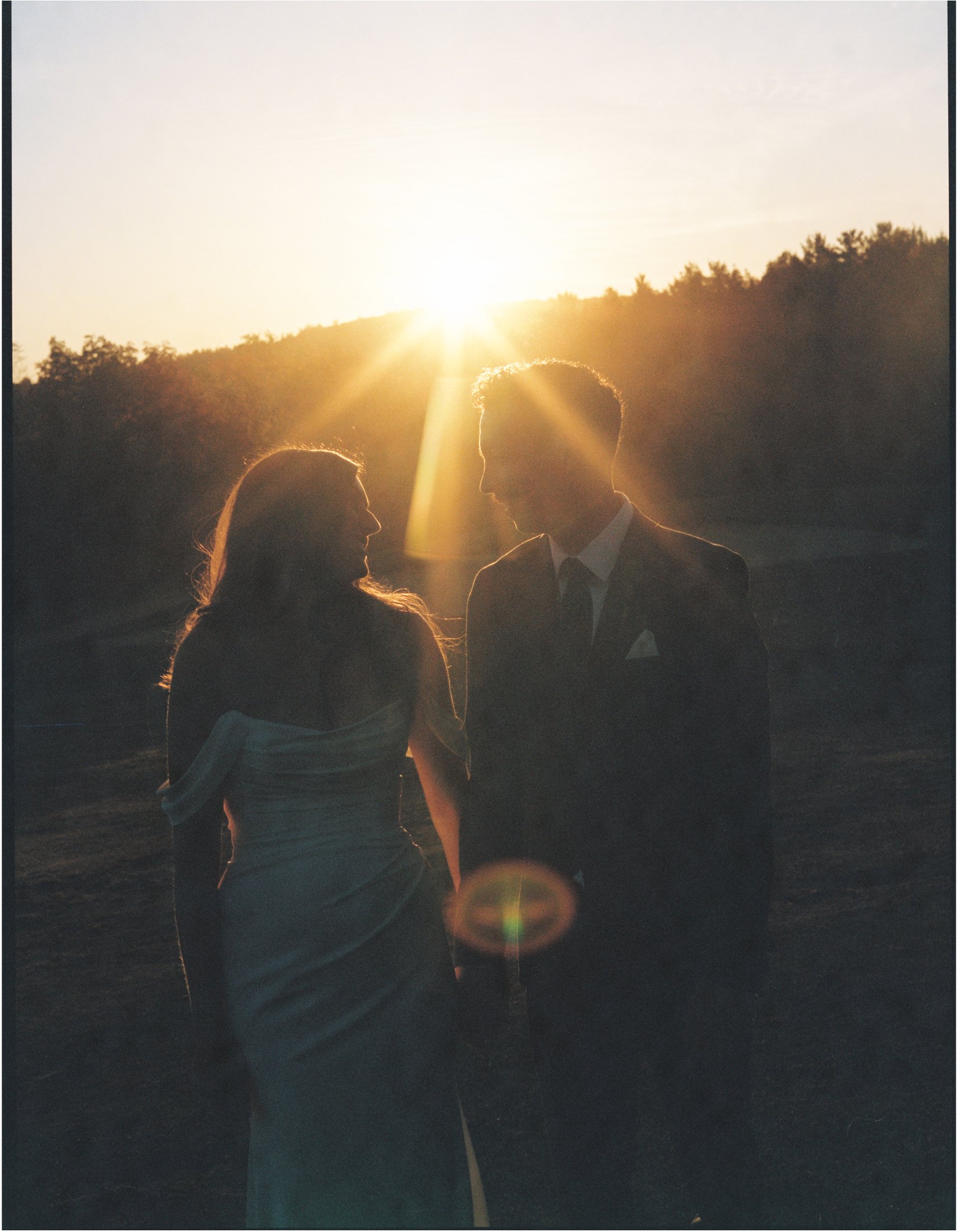 A couple dressed in formal attire holding hands at sunset outdoors with silhouettes and golden sunlight.