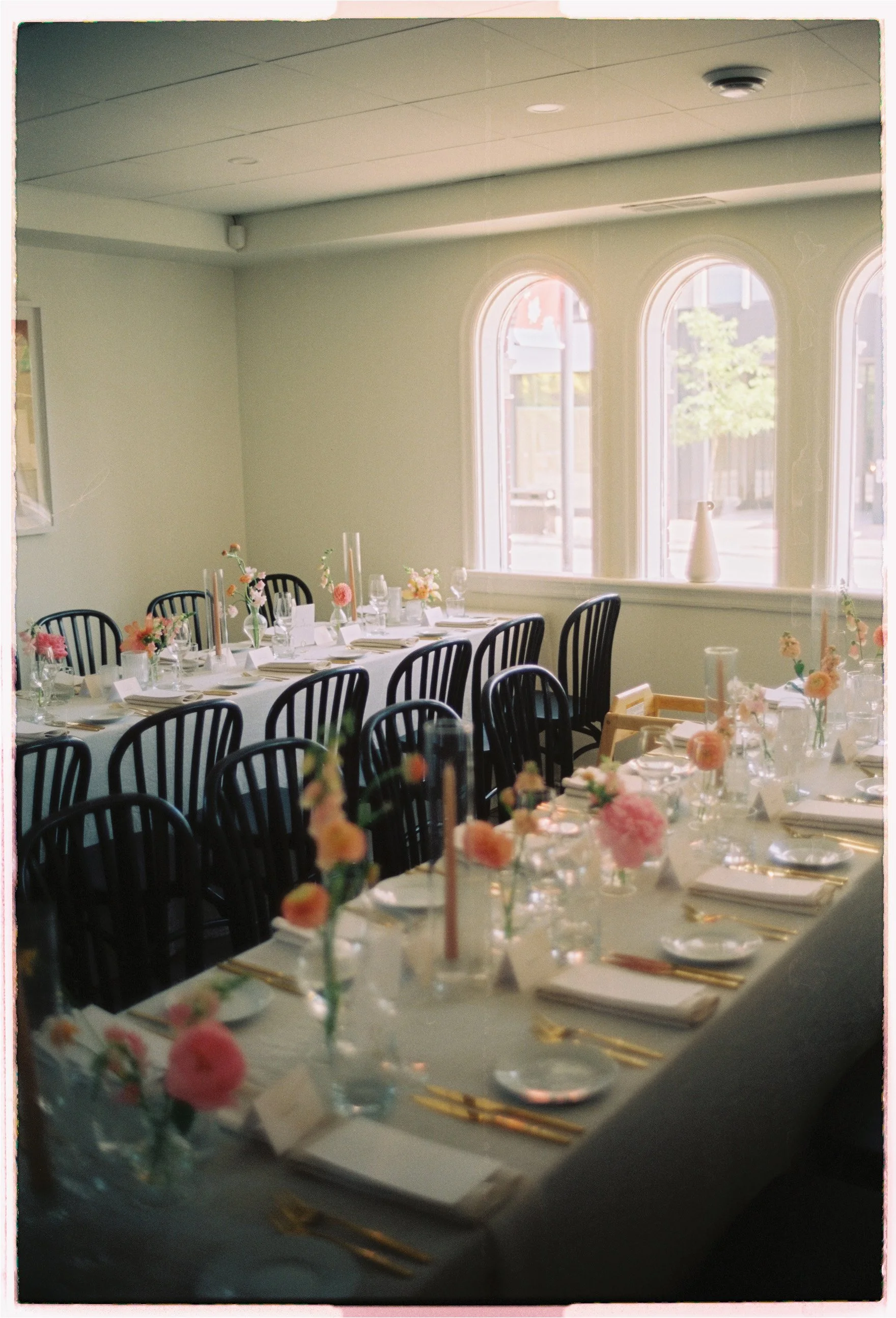 A decorated dining table set for an event with pink and peach flowers in glass vases, gold utensils, white plates, and napkins, in a room with large arched windows.